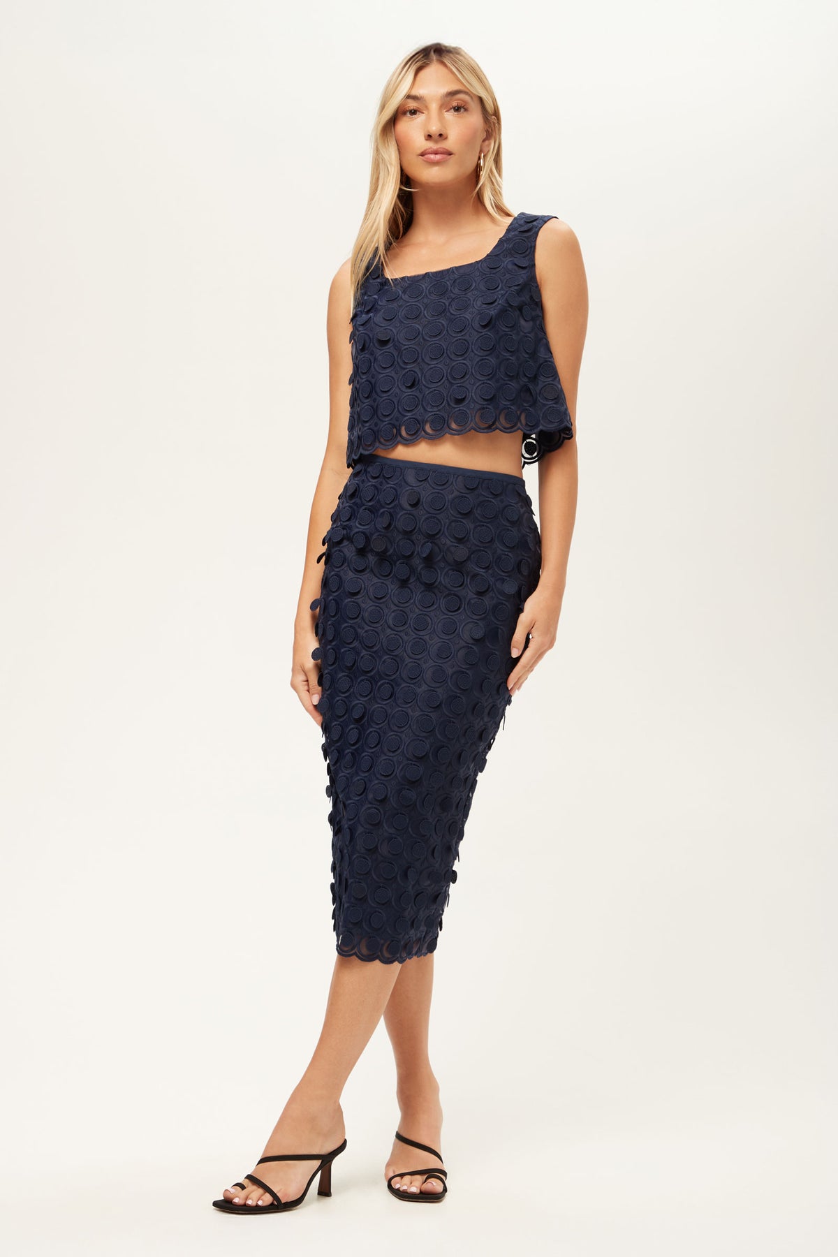 A woman wears the Dita 2 Midi Pencil Skirt - Agassi Embroidery, a navy blue high-waisted, knee-length skirt with a textured circular pattern, featuring a matching sleeveless crop top and black open-toe heels against a plain background.