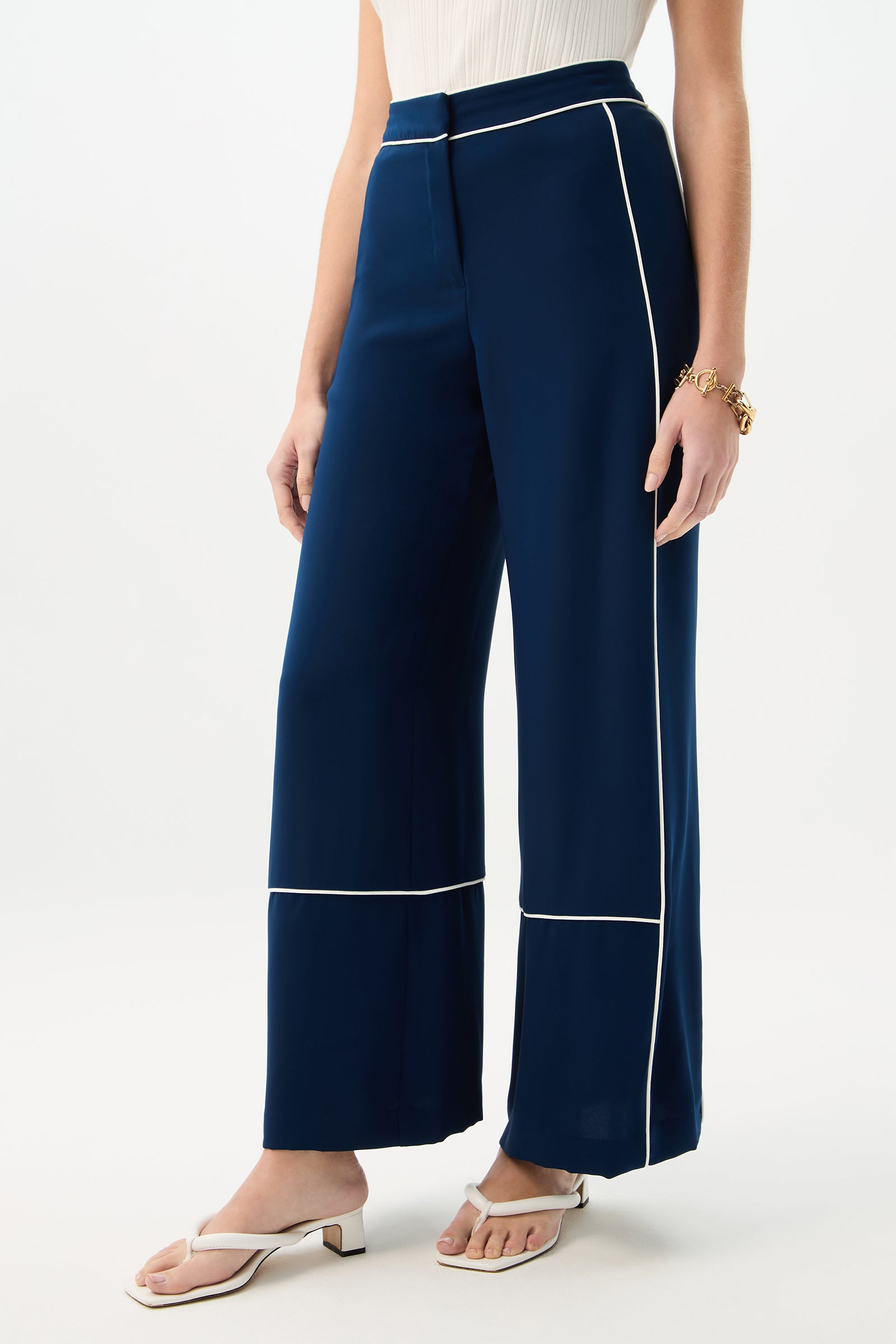 Wearing the Rivals Wide-Leg Pant - Double Georgette in navy with white piping, a sleeveless white top, white heeled sandals, and a gold bracelet, a person stands against a plain light background.