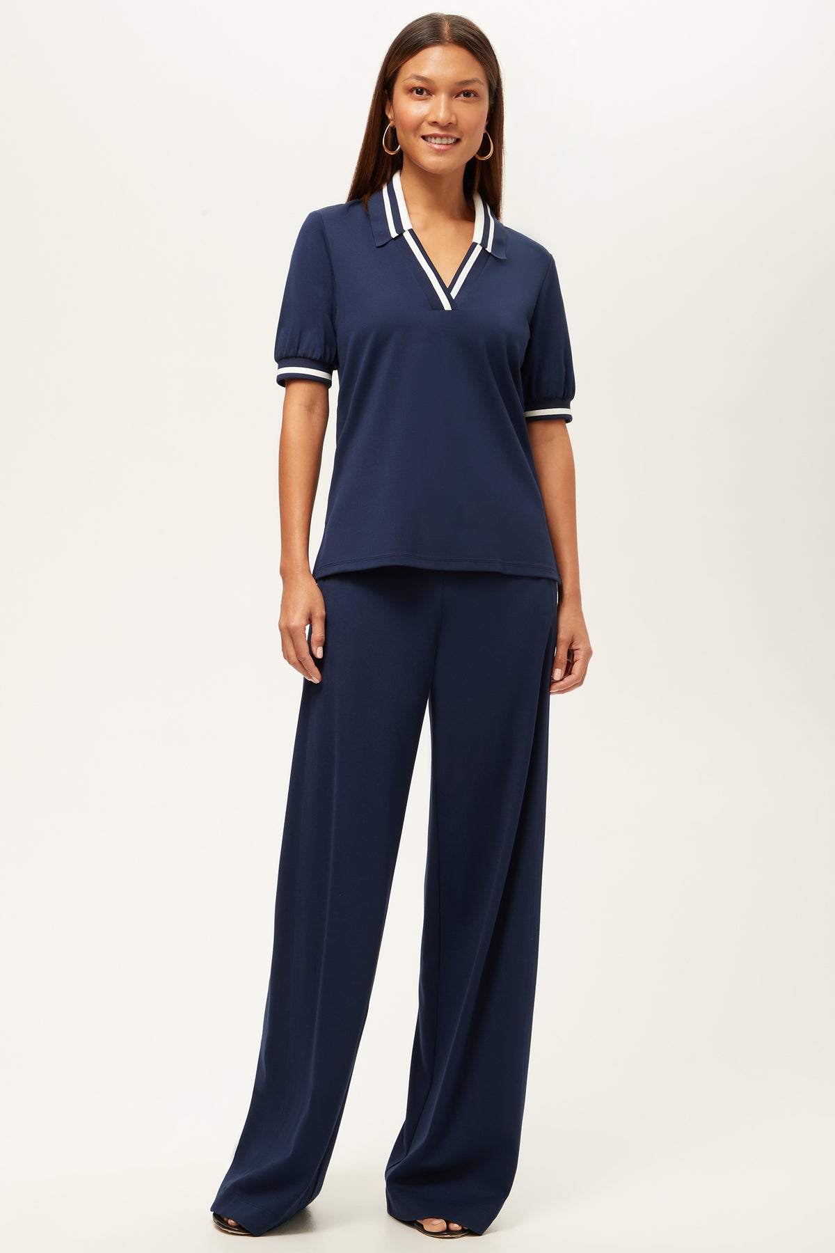 A woman with straight brown hair smiles in matching wide-leg navy pants, the Record Essential Wide-Leg Pant - Cozy Ponte, and a navy top with white striped collar and cuffs, posing against a plain white background.