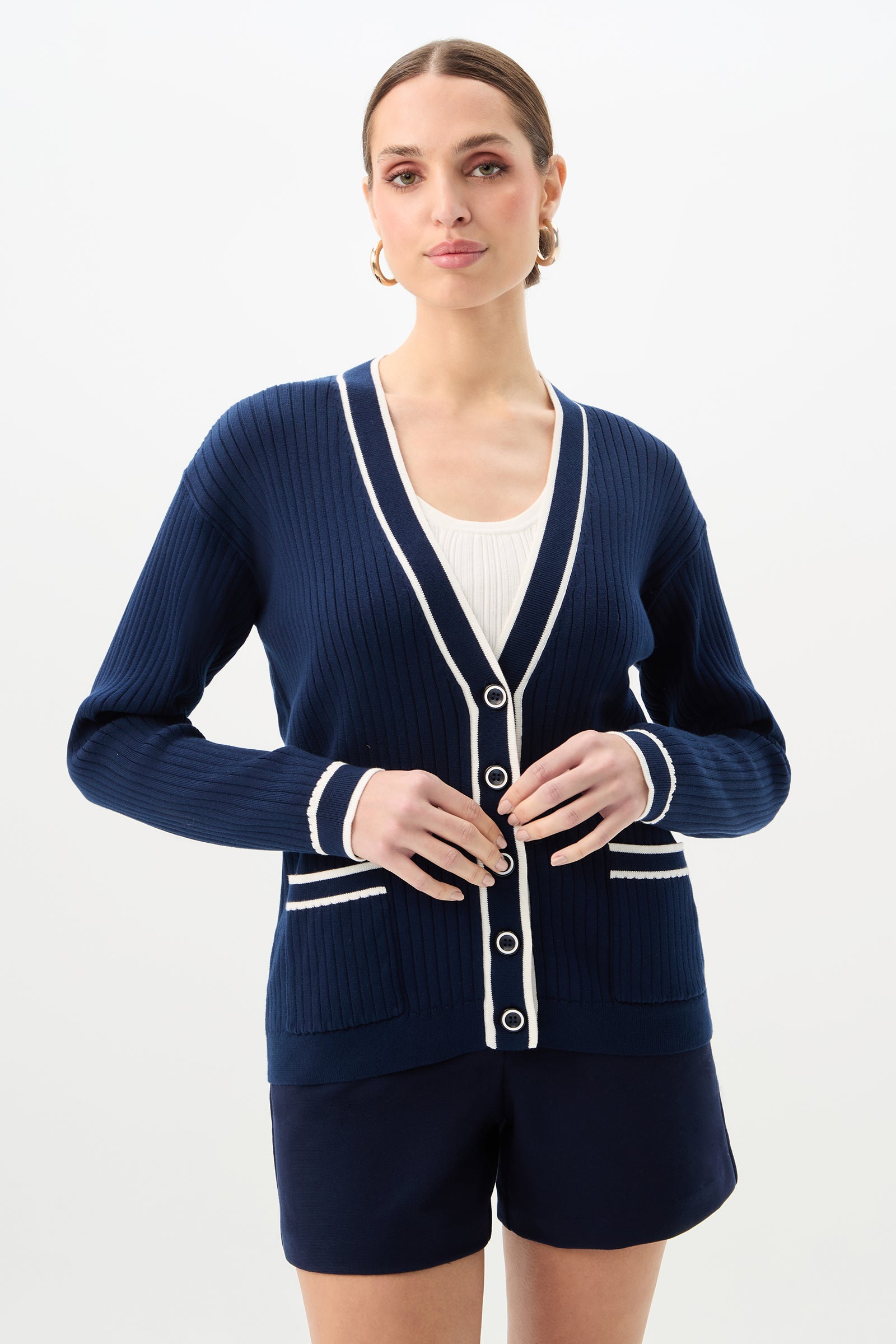 A woman wearing the Henin Contrast Trim V-Neck Knit Cardigan in Ink/Whitewash over a white top and navy shorts stands against a plain white background.