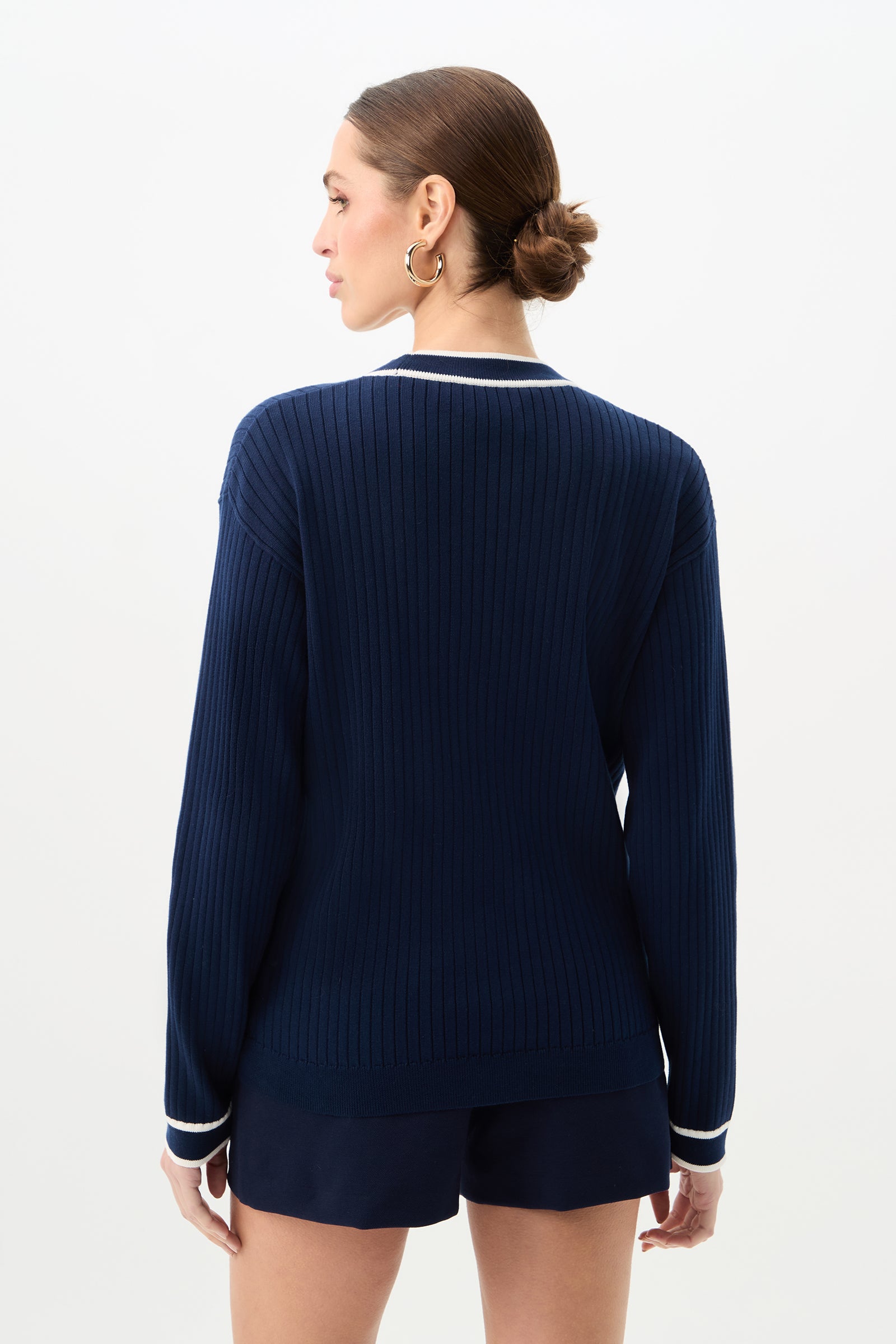 A woman with brown hair in a low bun models the Henin Contrast Trim V-Neck Knit Cardigan in navy blue, featuring white trim, paired with navy shorts. She is facing away from the camera against a plain white background.