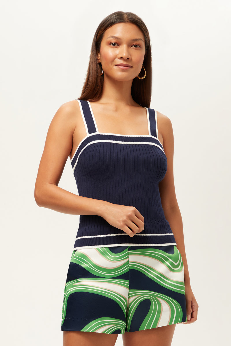 A woman with long straight hair smiles slightly while wearing the Break Point Contrast Trim Knit Cami Top in Ink/Whitewash and high-waisted shorts with a green, white, and navy abstract swirl pattern against a plain background.