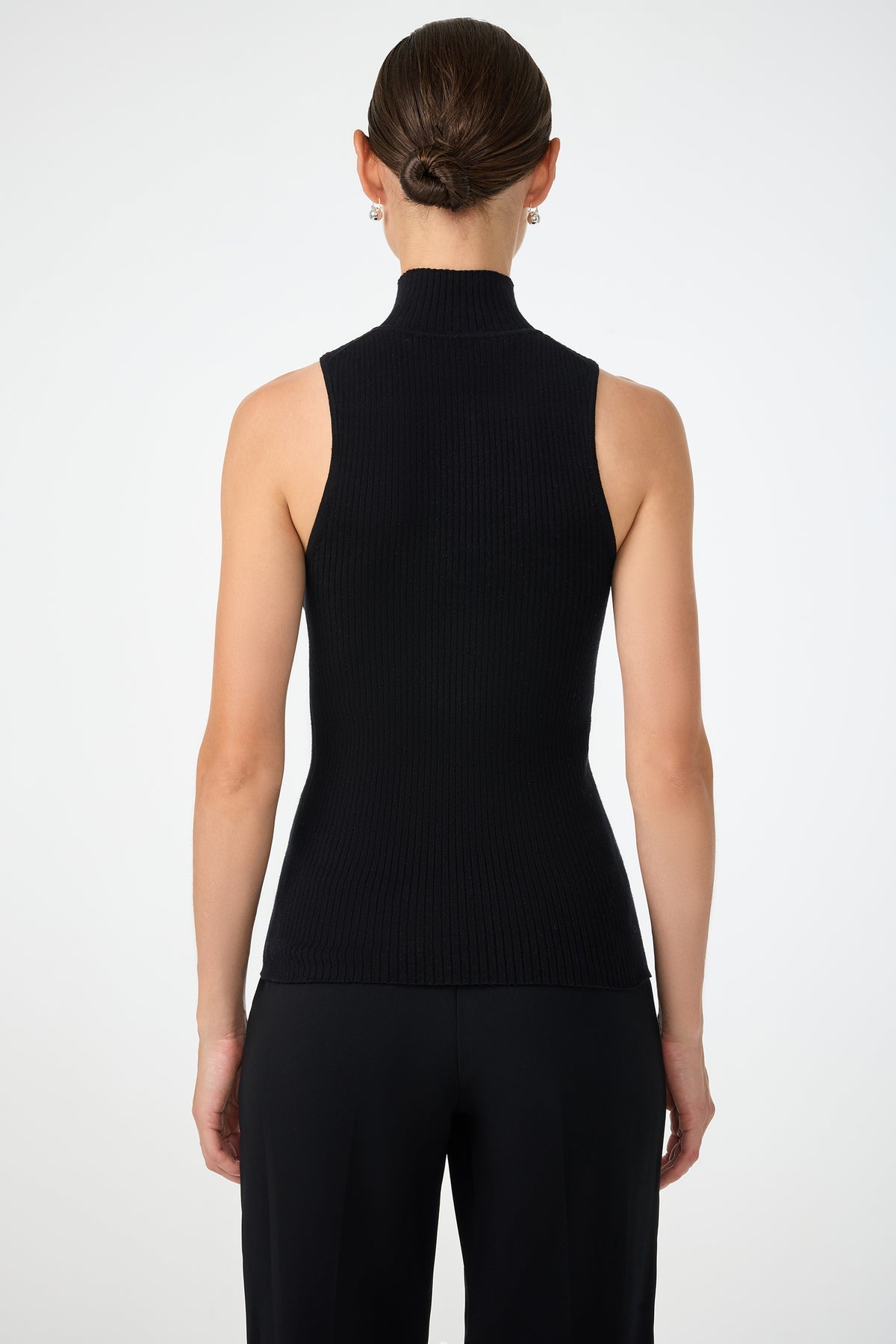 A woman with her hair tied back is shown from behind wearing the Westhampton Knit Pullover, a sleeveless black turtleneck top, and black pants against a plain white background.