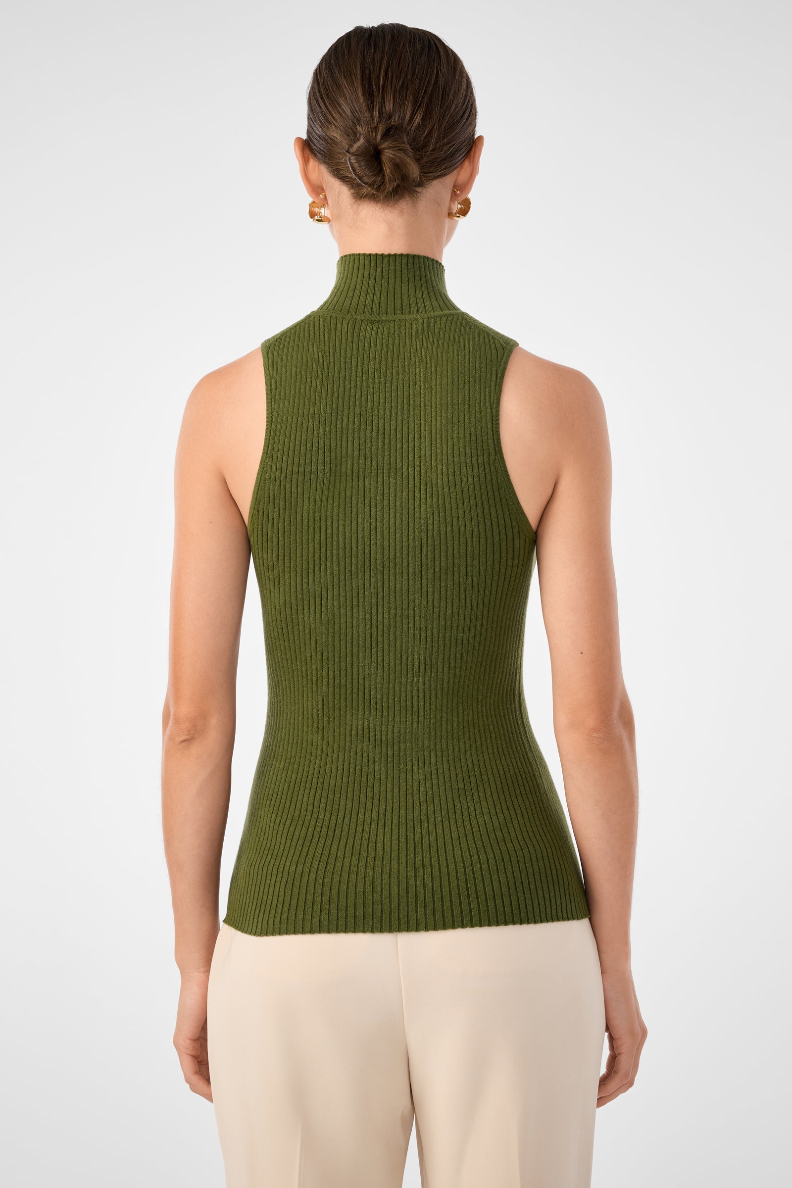 A woman faces away, wearing the Westhampton Knit Pullover—an olive green, sleeveless ribbed turtleneck—paired with light beige pants and gold hoop earrings. Her brown hair is styled in a low bun against a plain white background.