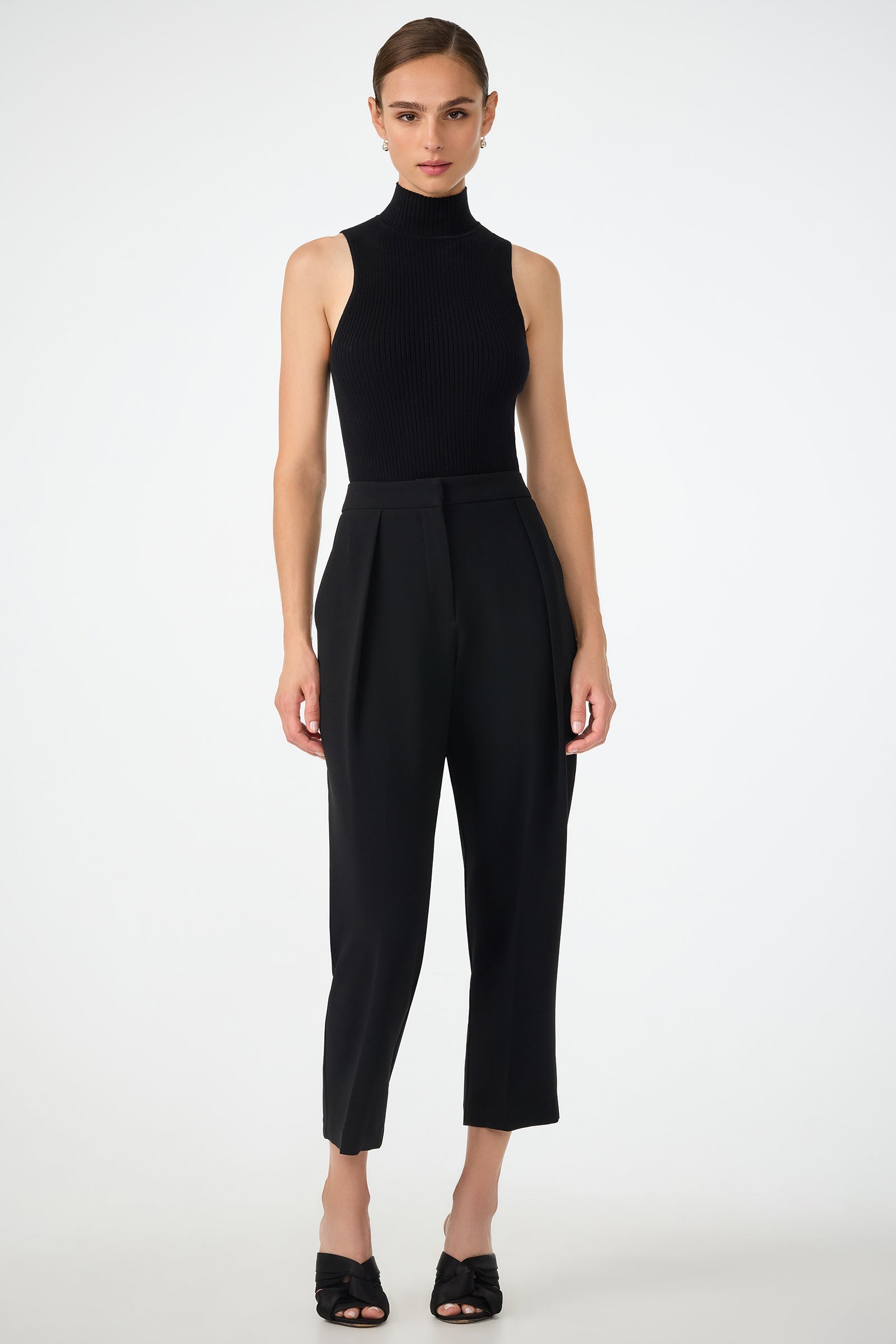 A woman stands against a plain white background, wearing the sleeveless black Westhampton Knit Pullover with high-waisted black tailored pants and open-toe heels. Her hair is pulled back and she has a neutral expression.
