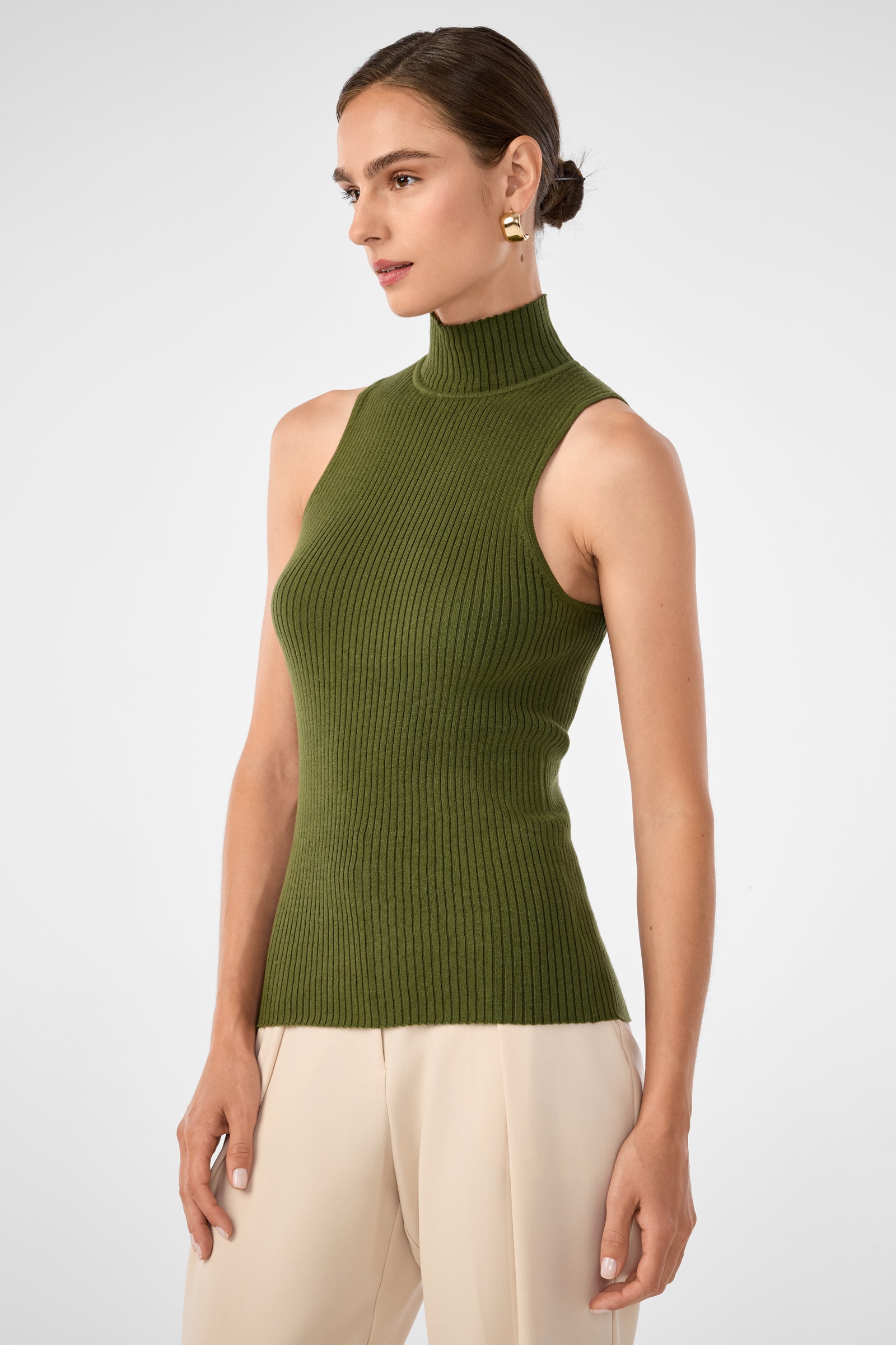 A woman wears the Westhampton Knit Pullover, a sleeveless ribbed olive green mock-neck top, paired with beige pants. She stands against a plain light background with her hair pulled back and gold earrings.