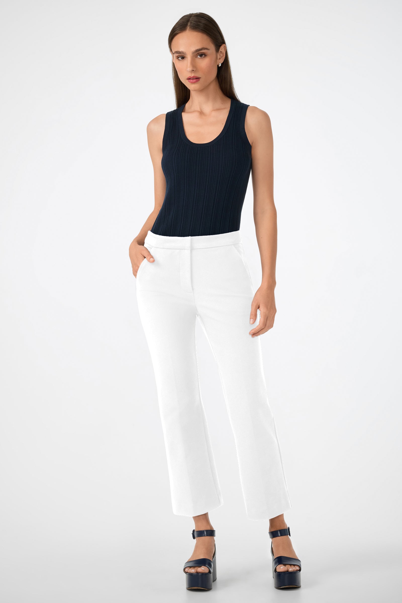 A woman with straight brown hair and a neutral expression stands against a plain background wearing Banshee Cropped Cuff Hem Pants, a fitted sleeveless black top, and navy blue platform sandals, with one hand in her pocket.