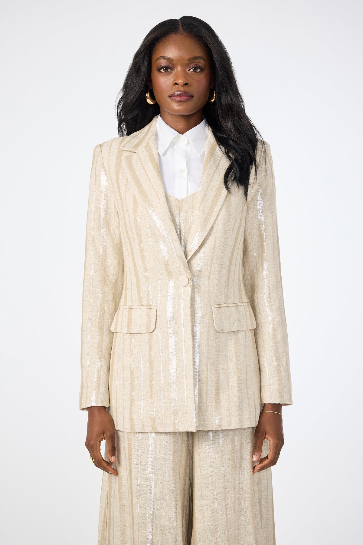 A woman faces forward in the Alternates 2 Blazer, a beige pinstripe suit with wide-leg trousers over a white collared shirt. She has long dark hair, gold earrings, and a neutral expression against a plain background.