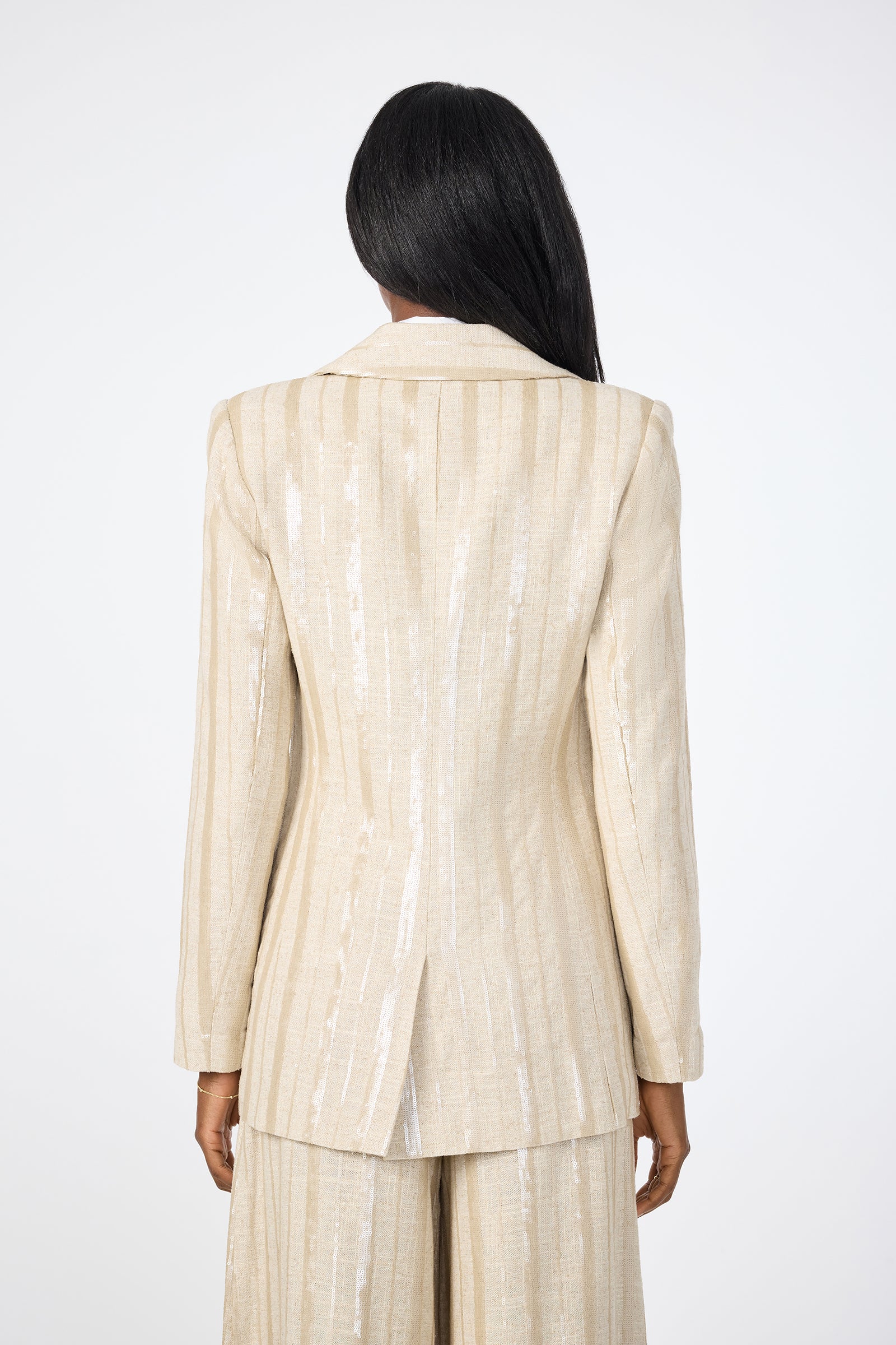 A person with long dark hair faces away, wearing the Alternates 2 Blazer—a light beige, shimmery pinstripe suit jacket with matching pants—set against a plain white background.