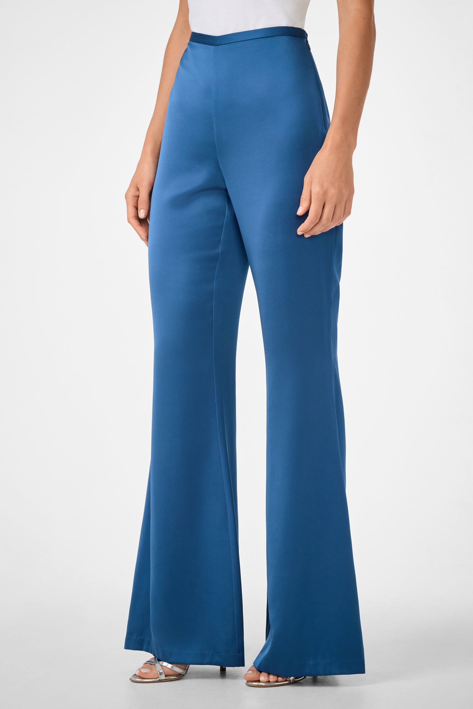 A person wearing Anmitsu Flared Pants—high-waisted, wide-leg blue satin trousers—with a white top and silver heeled sandals, standing against a plain light background.