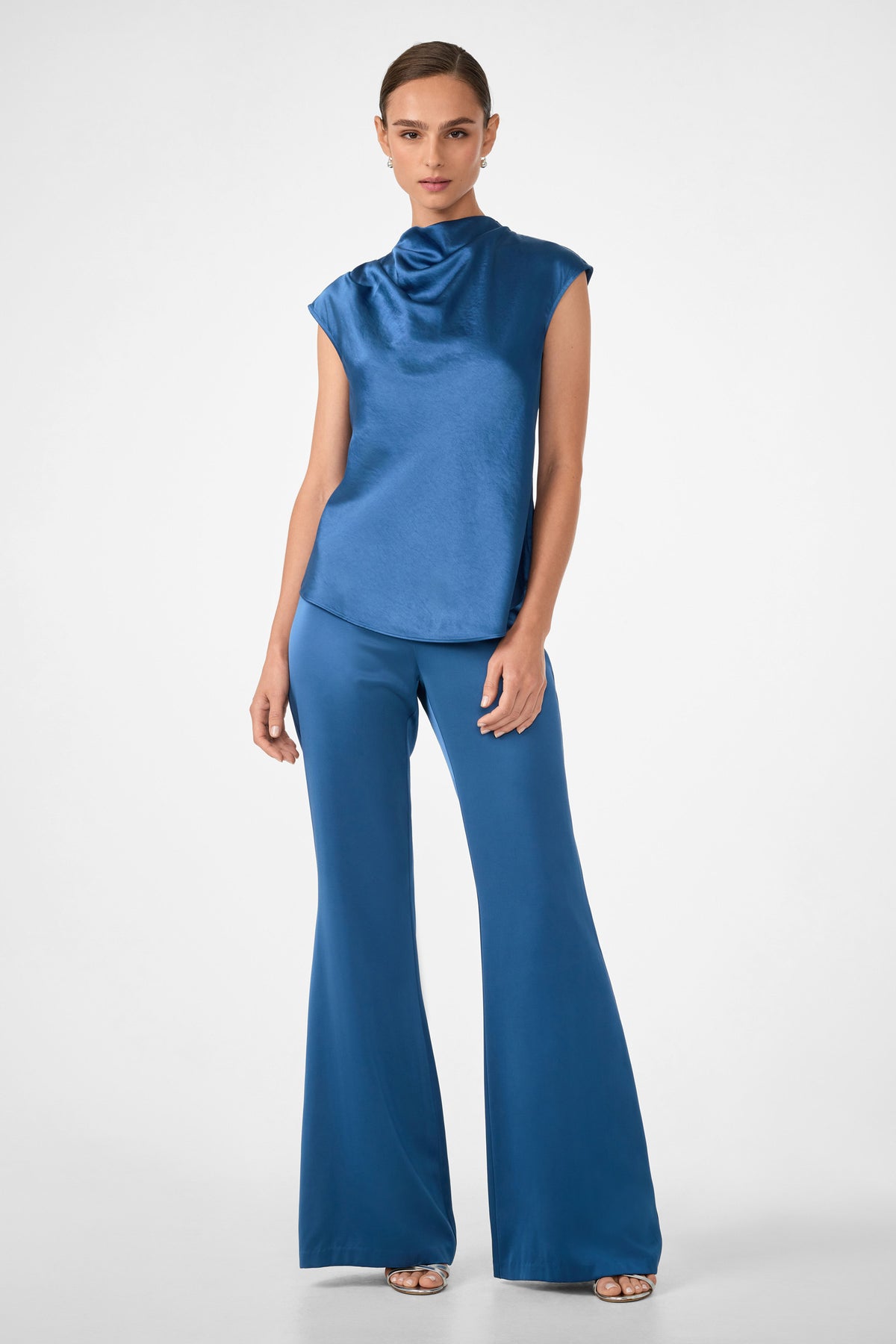 A woman poses against a plain background wearing shiny blue Anmitsu Flared Pants with a matching sleeveless, high-neck satin top and open-toe heels.