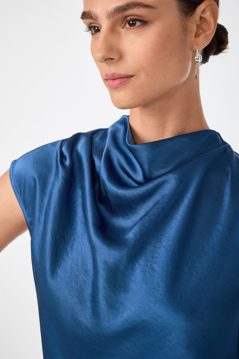 A woman in the Mikan Sleeveless Top, a shiny blue piece with a draped neckline, has her hair pulled back and wears small dangling earrings, gazing slightly to the side with a neutral expression.