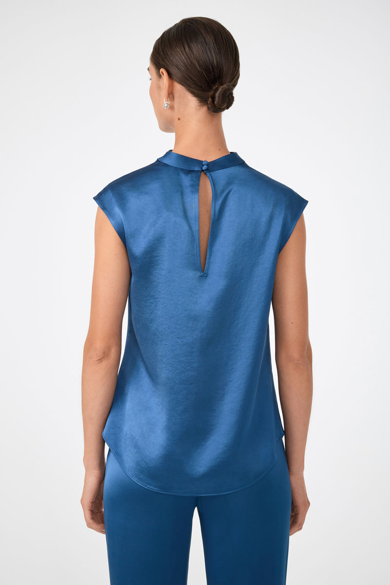 A woman with brown hair in a low bun is seen from the back, wearing the shiny blue Mikan Sleeveless Top with a keyhole detail and matching blue pants, set against a plain light background.