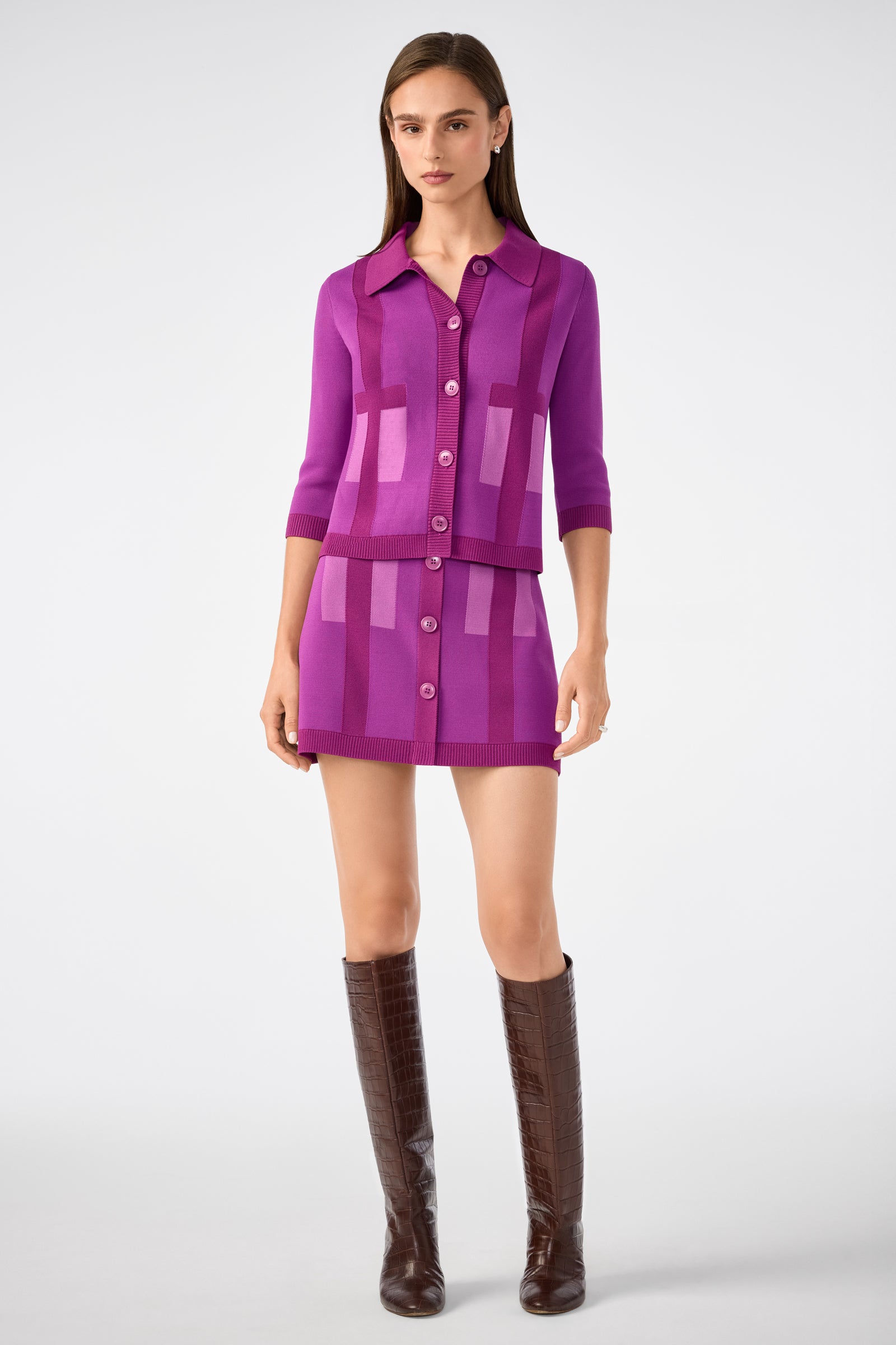A woman faces forward in a magenta Gaia Cardigan with matching geometric-patterned mini skirt, straight brown hair, and brown knee-high boots, against a plain white background.