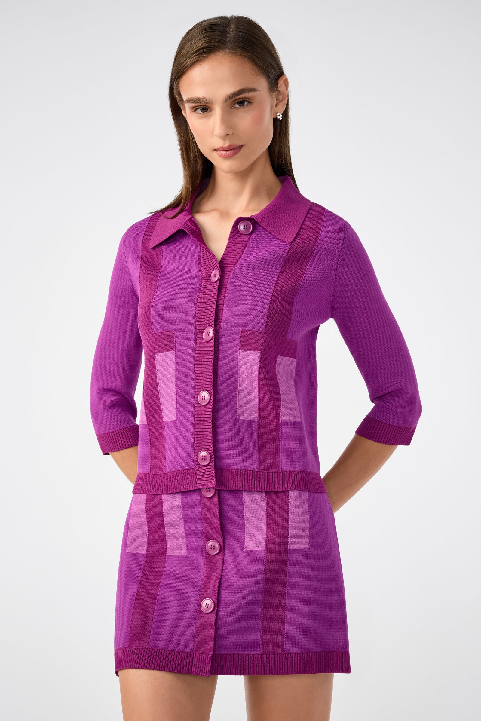 A woman wears the Gaia Cardigan, a vibrant purple button-up with three-quarter sleeves, paired with a matching geometric-patterned skirt, standing against a plain light background.