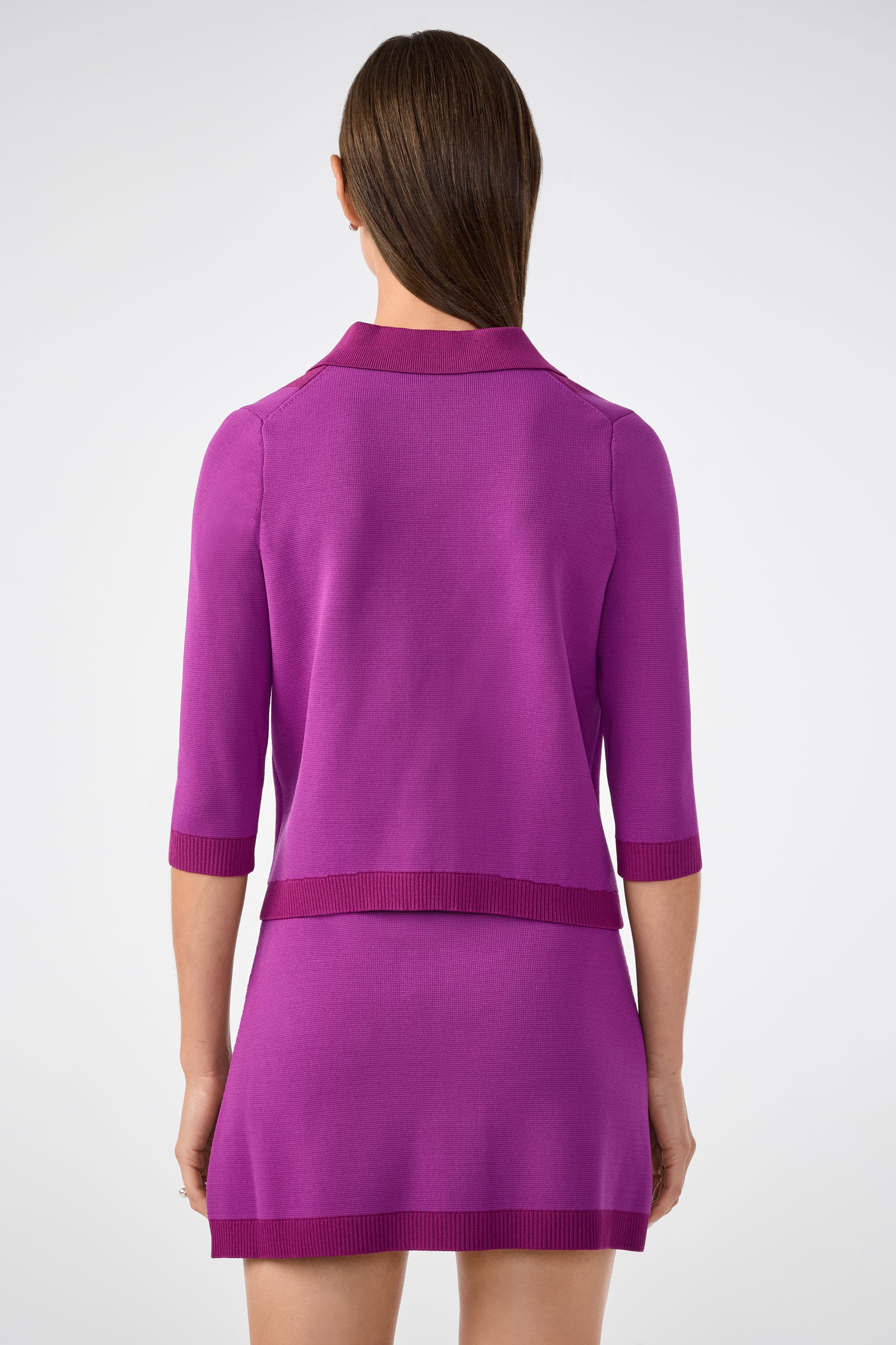 A woman with straight brown hair, seen from behind, wears the Gaia Cardigan in bright purple with a matching short skirt, standing against a plain light background.