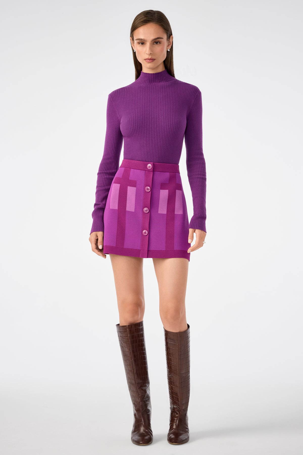 A woman poses against a white background in a fitted purple turtleneck, the Artemis Skirt—a high-waisted purple mini skirt with pink rectangular patterns and front buttons—and knee-high brown boots.