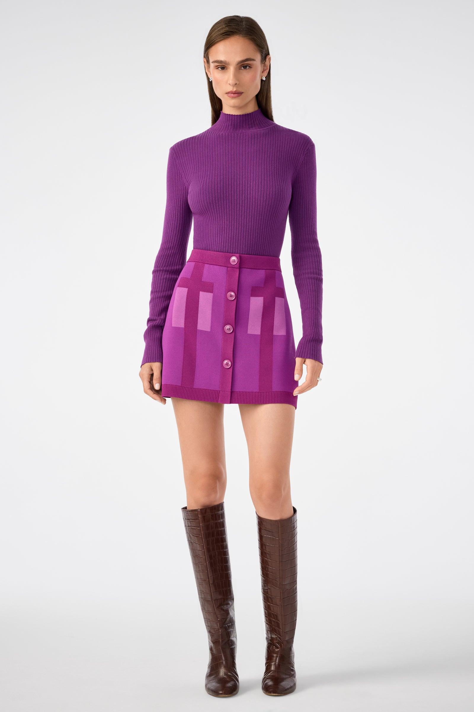 A woman poses against a white background in a fitted purple turtleneck, the Artemis Skirt—a high-waisted purple mini skirt with pink rectangular patterns and front buttons—and knee-high brown boots.