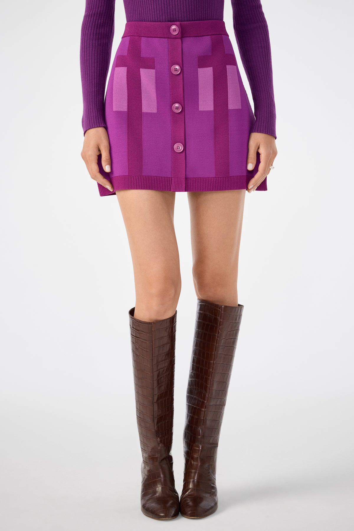 A person wears the Artemis Skirt—a purple and pink geometric-patterned mini skirt with buttons—paired with a purple long-sleeve top and knee-high brown boots, standing against a plain white background.