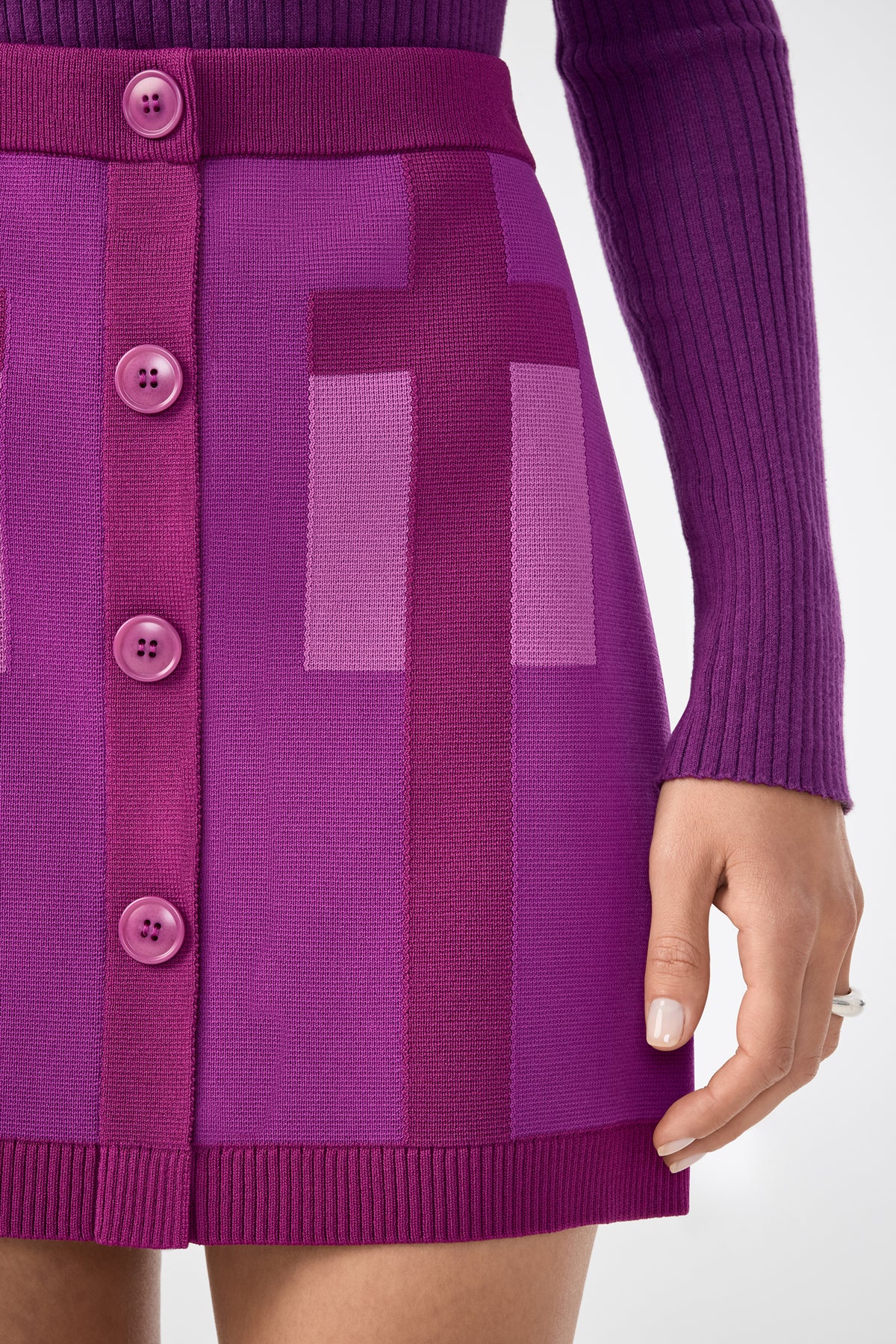 A person wearing the Artemis Skirt—a vibrant purple knit skirt with large front buttons and geometric patterns—paired with a ribbed long-sleeve top. Only the lower torso and one hand are visible.