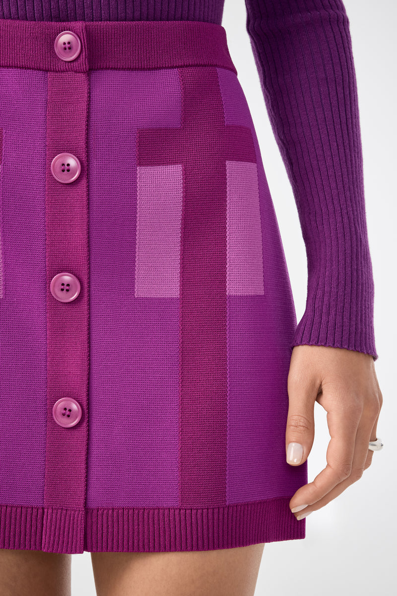 A person wearing the Artemis Skirt—a vibrant purple knit skirt with large front buttons and geometric patterns—paired with a ribbed long-sleeve top. Only the lower torso and one hand are visible.