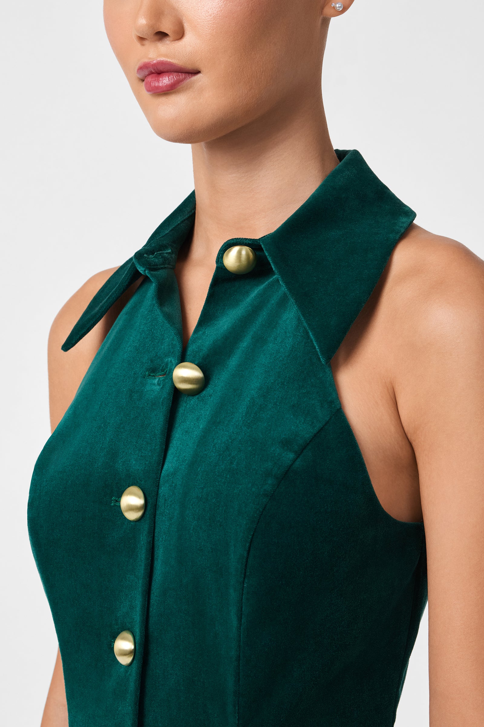 A woman models the Safina Dress, a sleeveless dark green velvet dress with a collar and large gold buttons, shown from shoulders to mid-torso against a plain background.