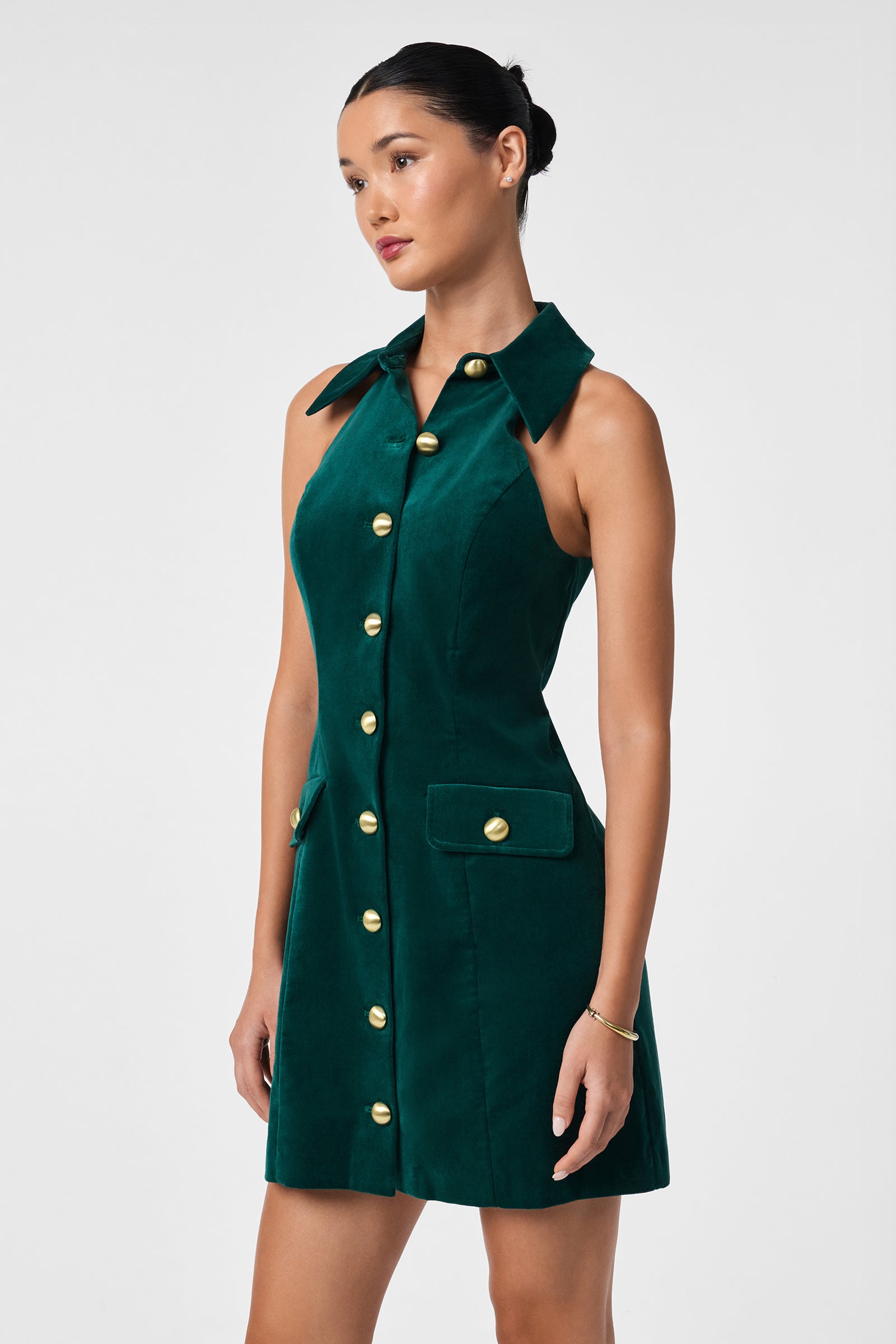 A woman wearing the Safina Dress, a sleeveless dark green velvet style with a wide collar, gold front buttons, and pocket flaps, gazes to the side. Her hair is styled in a neat bun and she accessorizes with a bracelet.