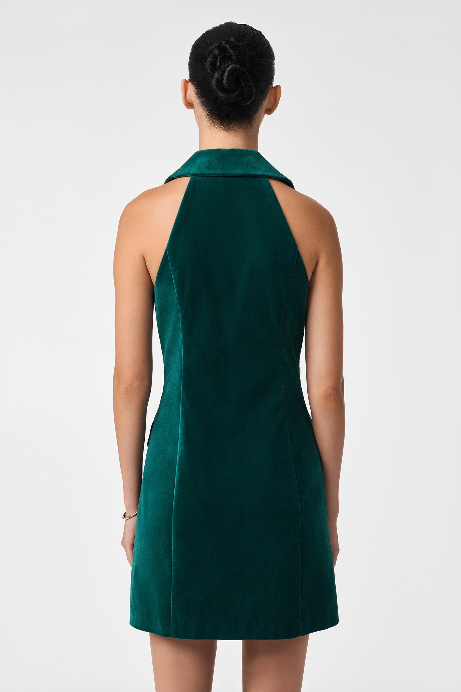 A person with dark hair in a bun is seen from the back wearing the sleeveless Safina Dress, an emerald green velvet design with a halter neckline, set against a plain light background.