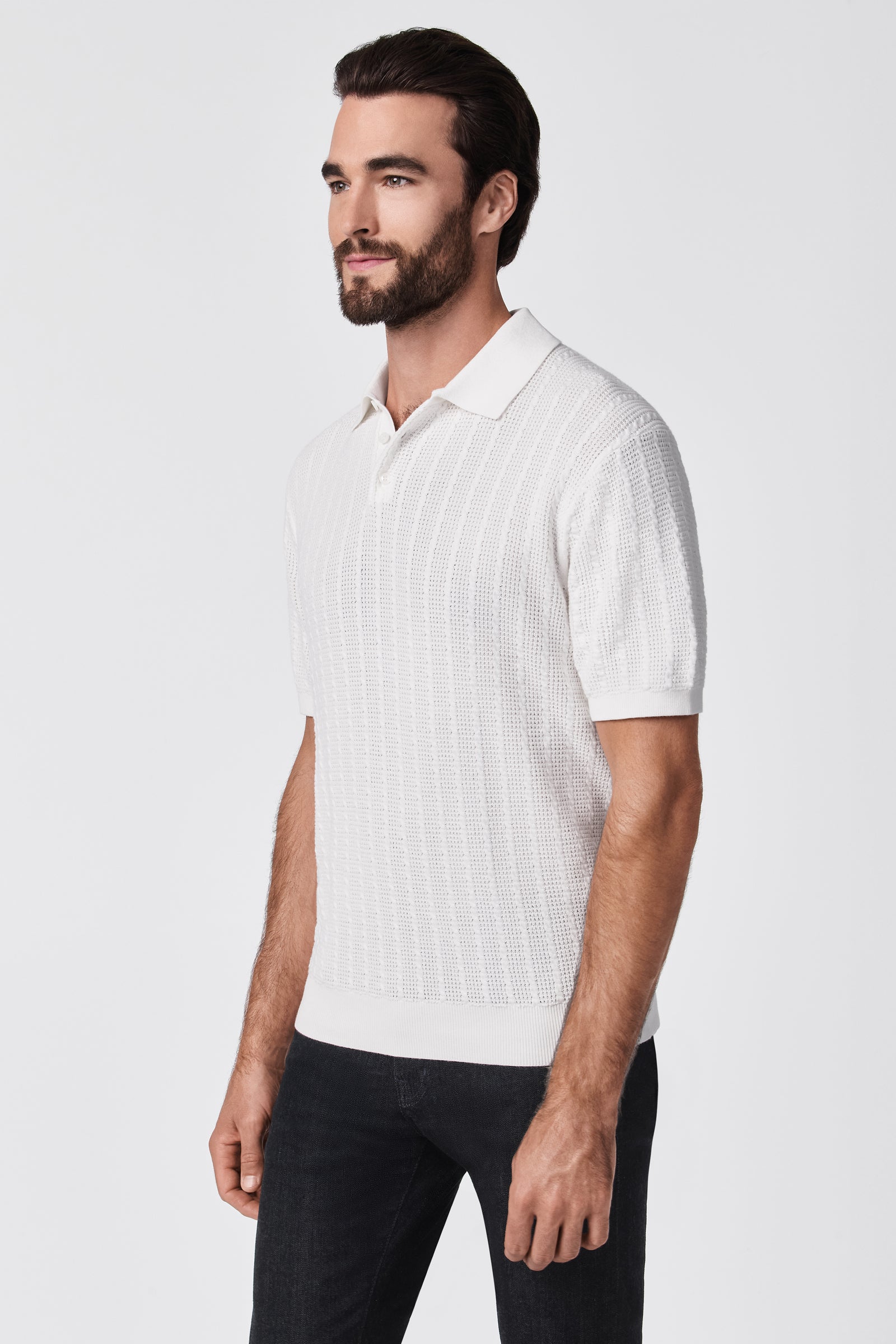 A man with dark hair and a beard is wearing the Asher Short-Sleeve Polo in white with black jeans, standing against a plain white background.