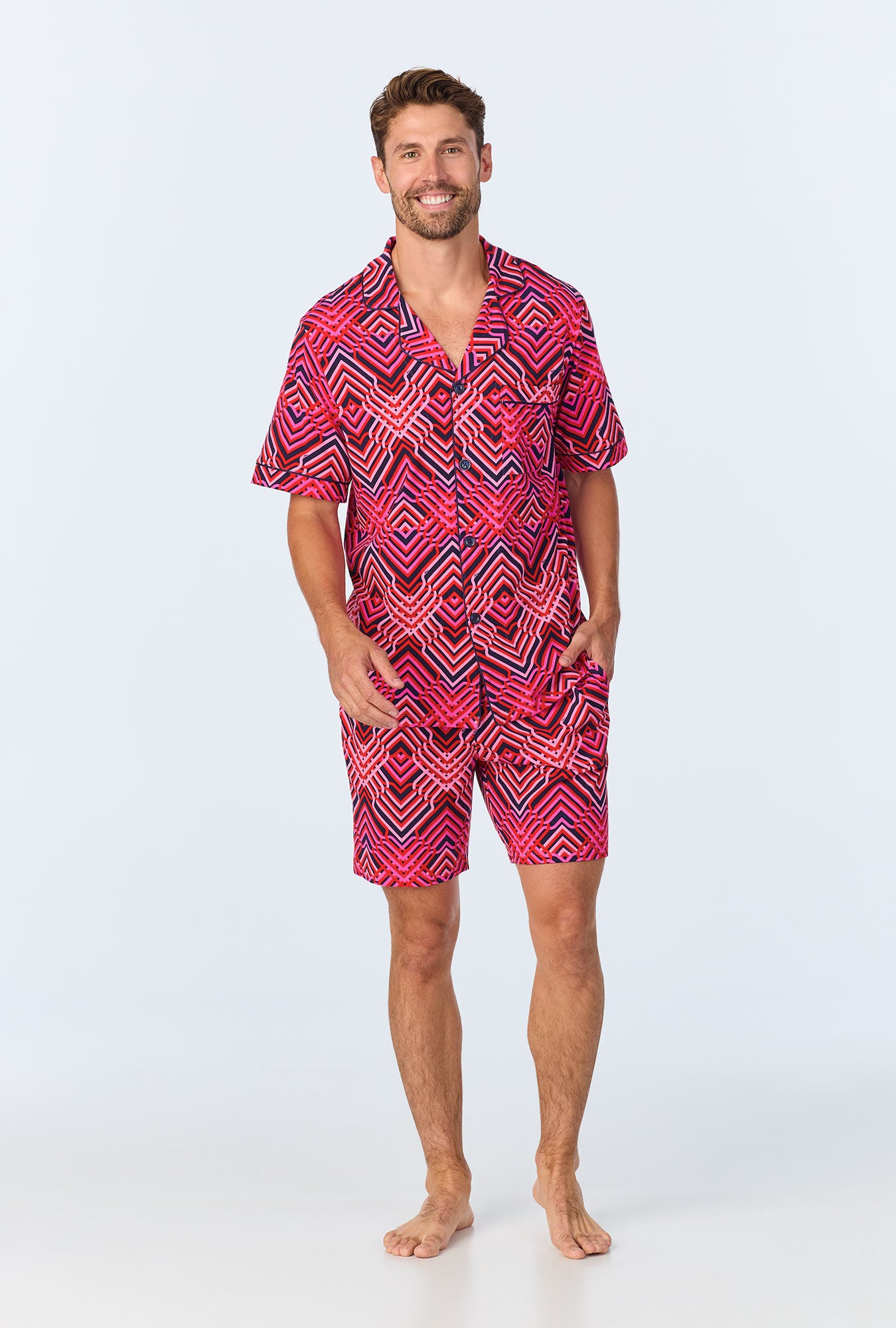 A man stands barefoot, smiling, wearing the Geo Trellis Mens Short-Sleeve Boxer Short Jersey PJ Set in multi, featuring a bright pink and black geometric pattern on a light background.