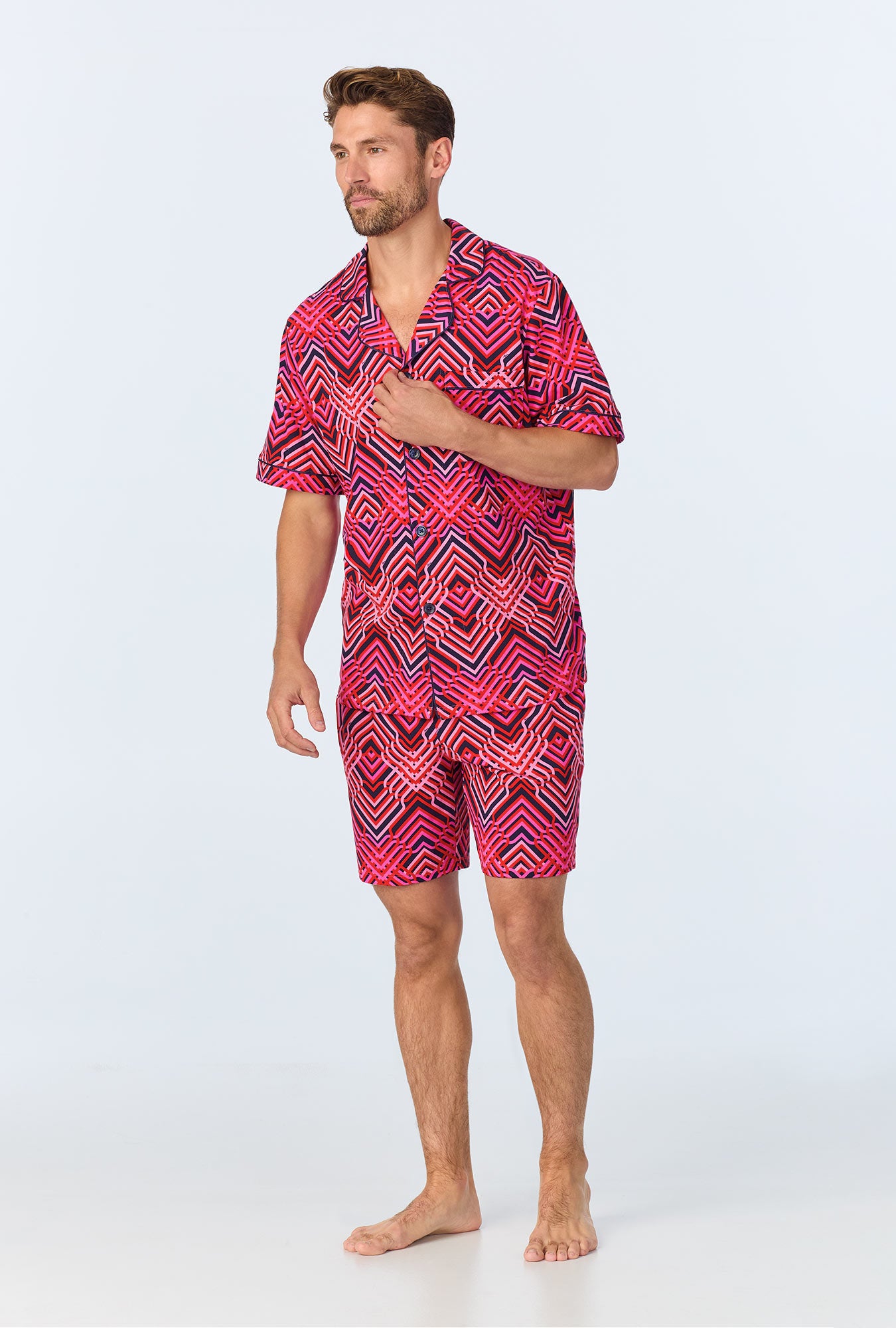 A barefoot man stands against a light background, wearing the Geo Trellis Mens Short-Sleeve Boxer Short Jersey PJ Set, which features a matching button-up shirt and shorts with a bold red, pink, and black geometric pattern.