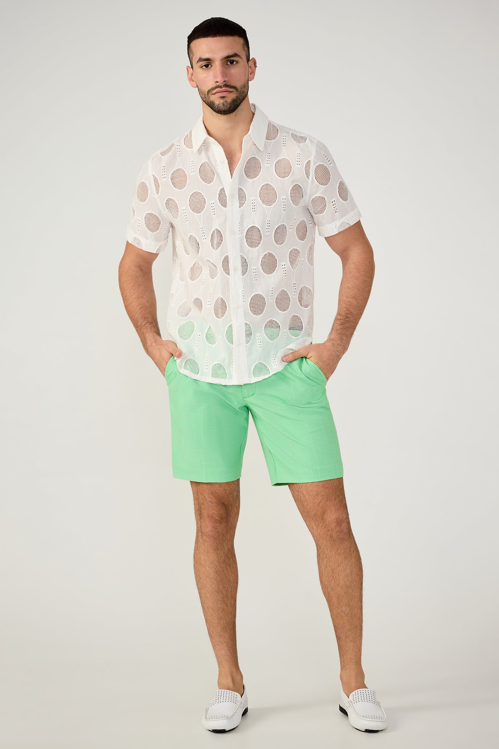 A man stands facing forward with hands in his pockets, wearing a white short-sleeve sheer shirt with large circular cutouts, the Travis Short - Game Time Glen Plaid in mint green, and white slip-on shoes. He has a neutral expression.