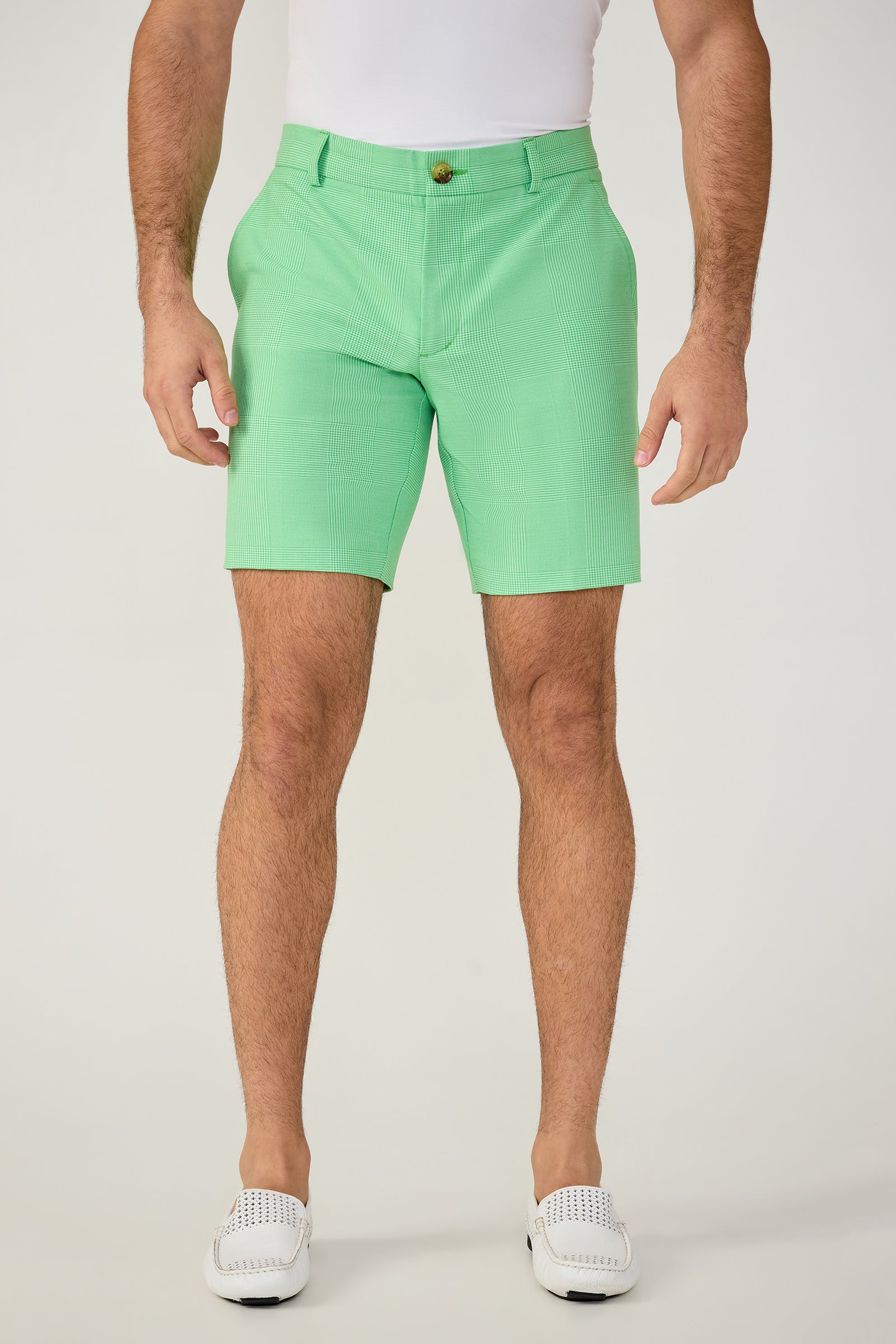 A man stands against a plain light background, wearing a white shirt, Travis Short - Game Time Glen Plaid in Grass Court Green/Whitewash, and white slip-on shoes. The image shows him cropped from shoulders to feet.
