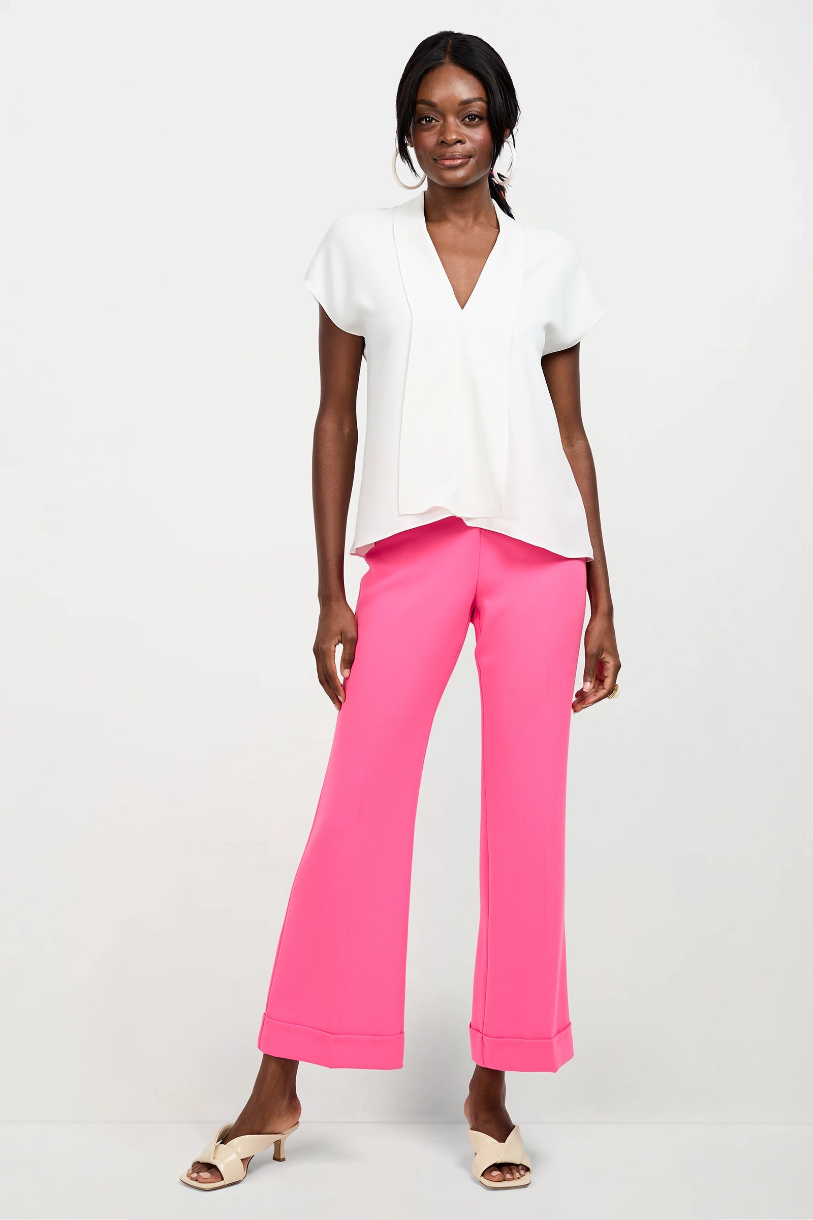 A woman stands against a white background wearing the Turin Slash Tie Top in Whitewash and bright pink wide-leg pants. She completes her look with beige open-toe heeled sandals and a soft smile.