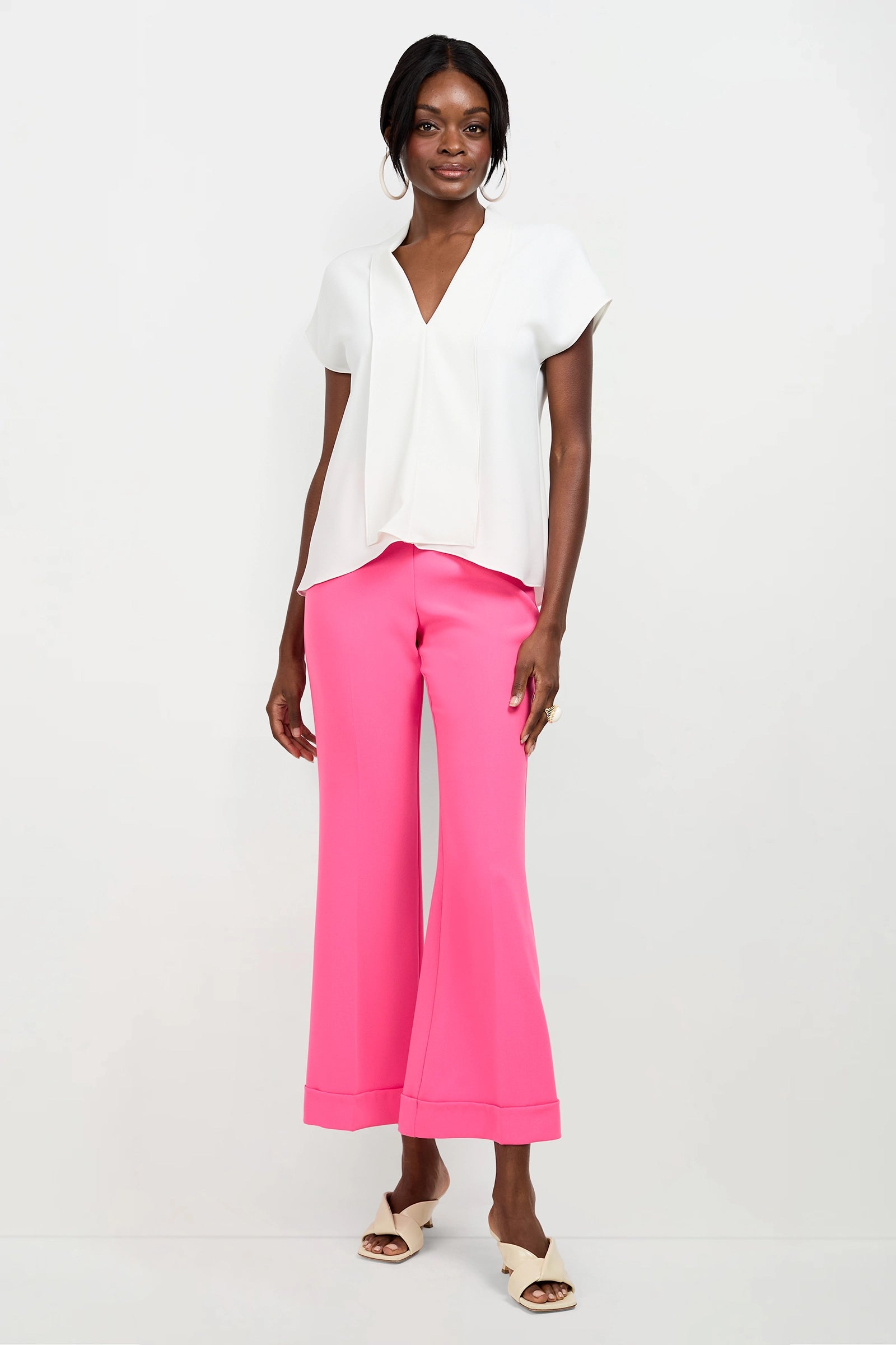 A woman stands against a white background wearing the Turin Slash Tie Top, bright pink wide-leg pants, white heeled sandals, and large hoop earrings. She smiles with her hands by her sides.