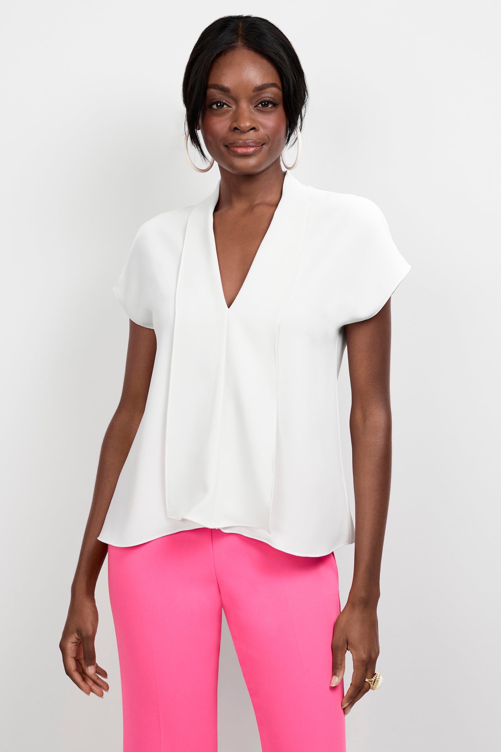 A woman models the Turin Slash Tie Top, a short-sleeve white V-neck blouse, paired with bright pink trousers. She has dark hair styled back, wears large hoop earrings, and smiles slightly against a white background.