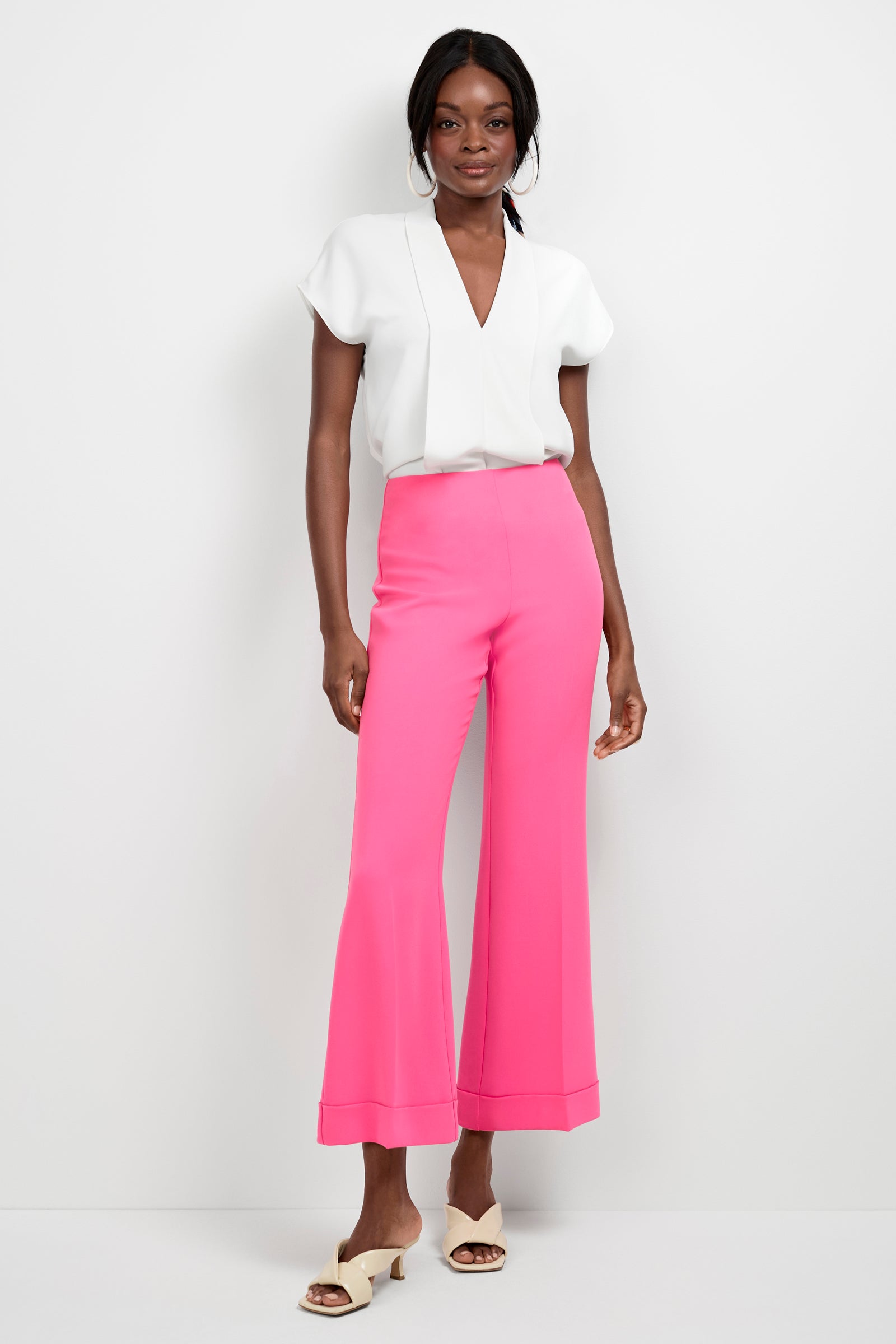A woman stands against a white wall, smiling and wearing high-waisted Loren Cropped Kick-Flare Pants in bright pink, a short-sleeve white blouse, and light-colored open-toe heels with one hand relaxed by her side.