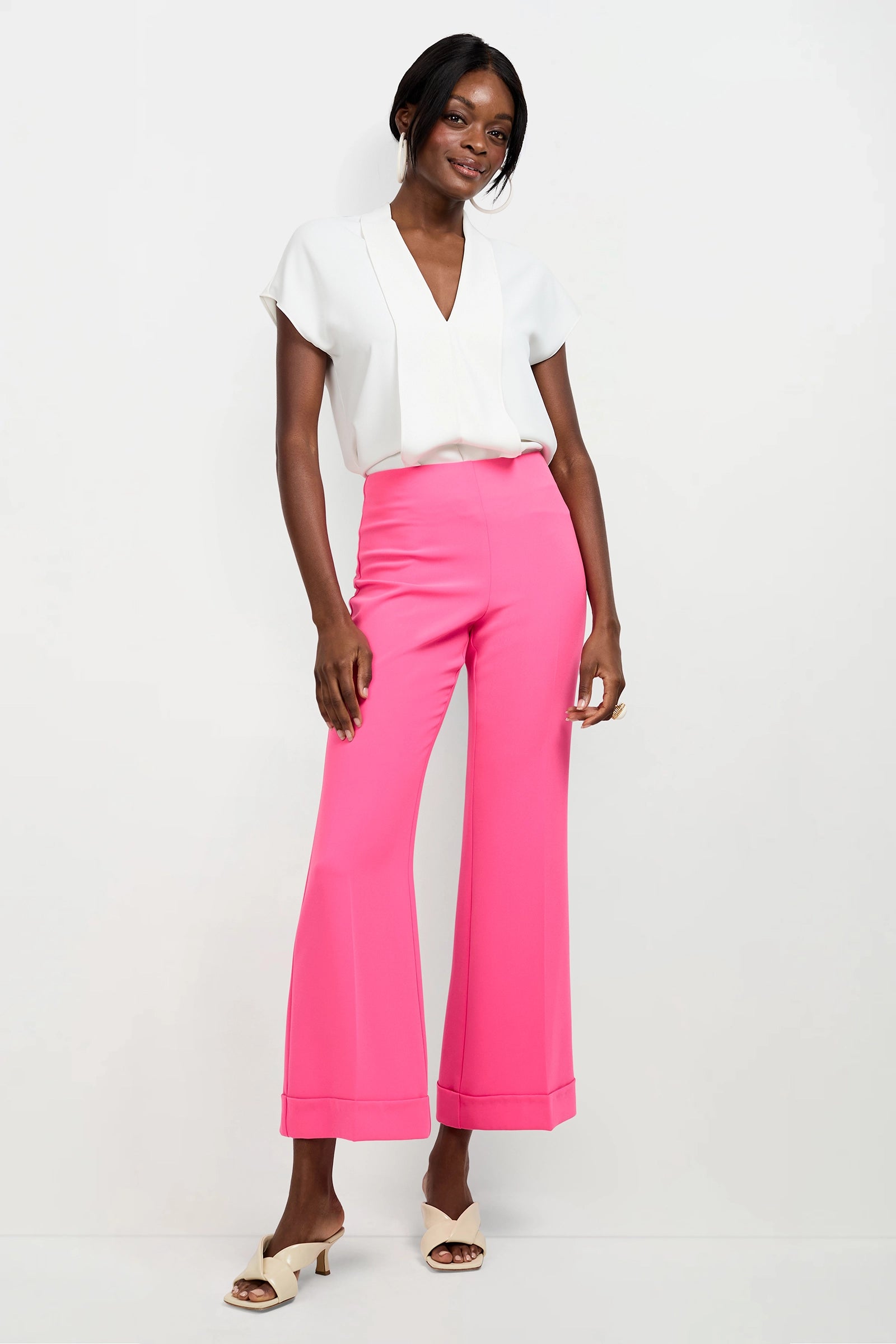 A woman stands against a white background, smiling with one hand on her hip, wearing the Turin Slash Tie Top, high-waisted bright pink wide-leg pants, and open-toe beige heels.