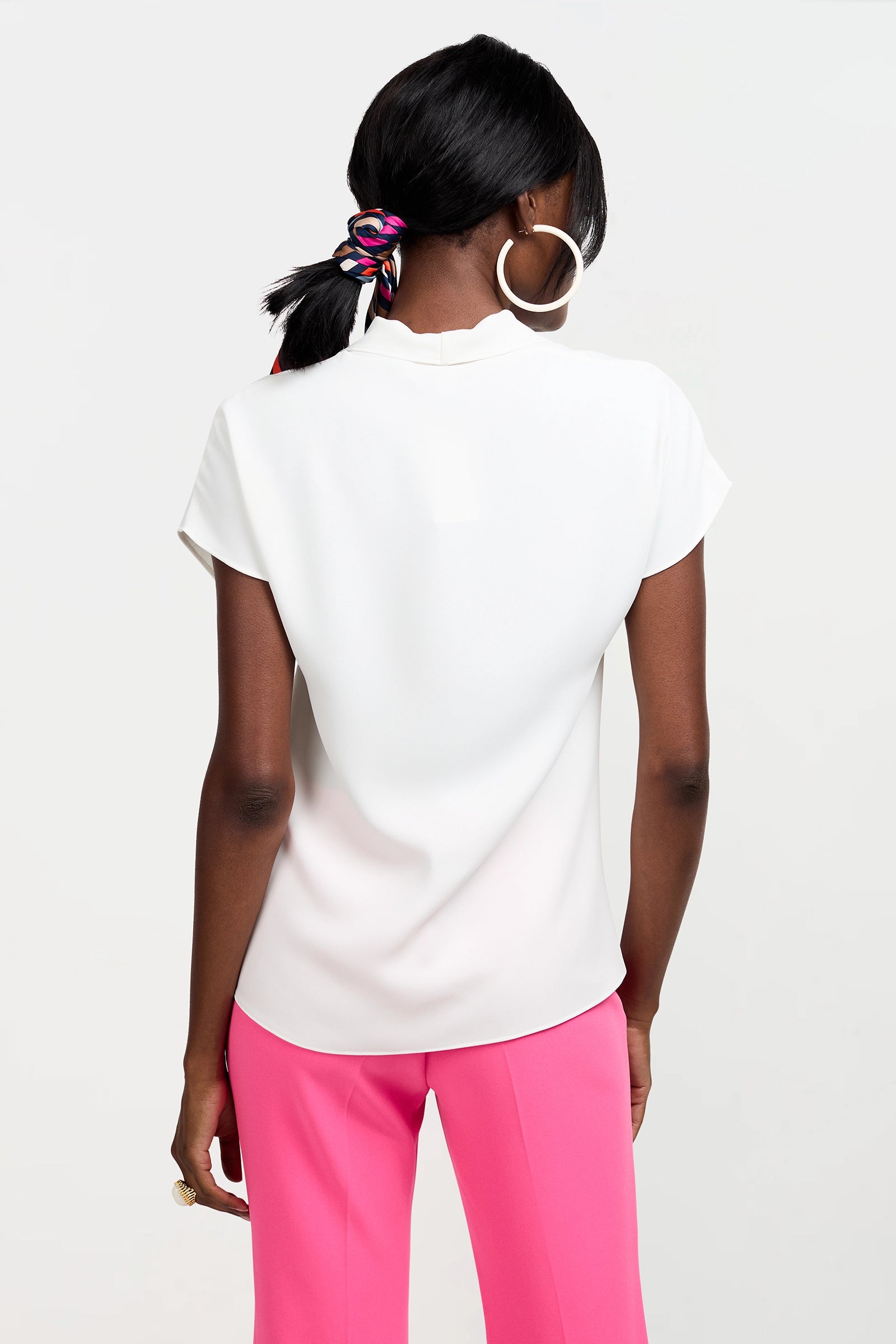 A woman with dark hair in a ponytail, wearing a colorful scrunchie, large hoop earrings, and the white Turin Slash Tie Top, stands against a light background, shown from the back.