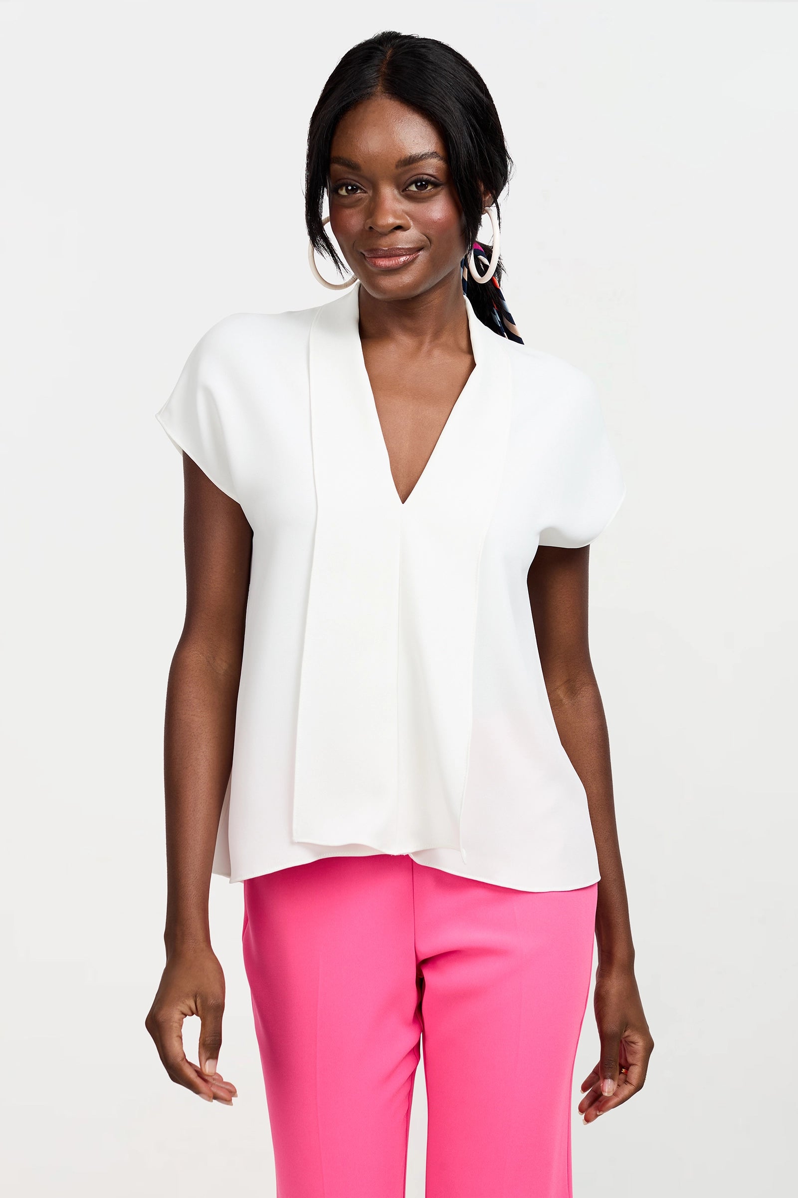 A woman stands against a plain white background, smiling and wearing the Turin Slash Tie Top—a short-sleeved white blouse with a V-neck—paired with bright pink pants, one hand relaxed at her side.