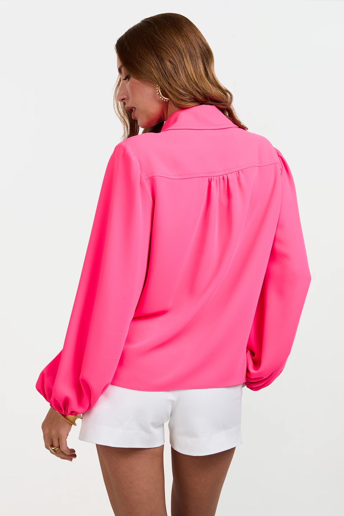 Woman with long brown hair wears the Lambro Peasant Top, a bright pink long-sleeve blouse, paired with white shorts. She stands with her back to the camera against a plain white background.