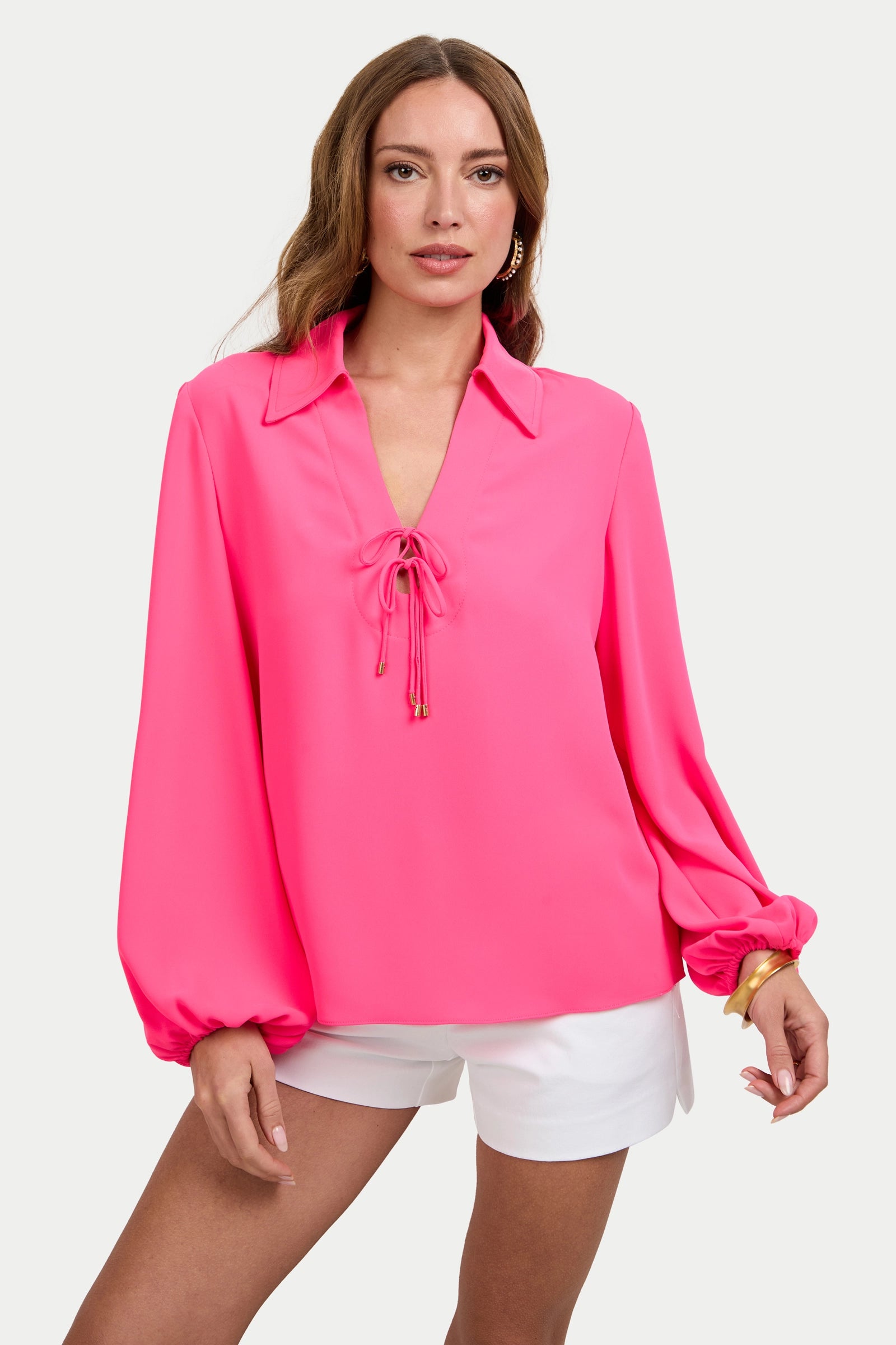 A woman wears the Lambro Peasant Top, a bright pink long-sleeve blouse with a lace-up neckline, paired with white shorts and standing against a plain light background.