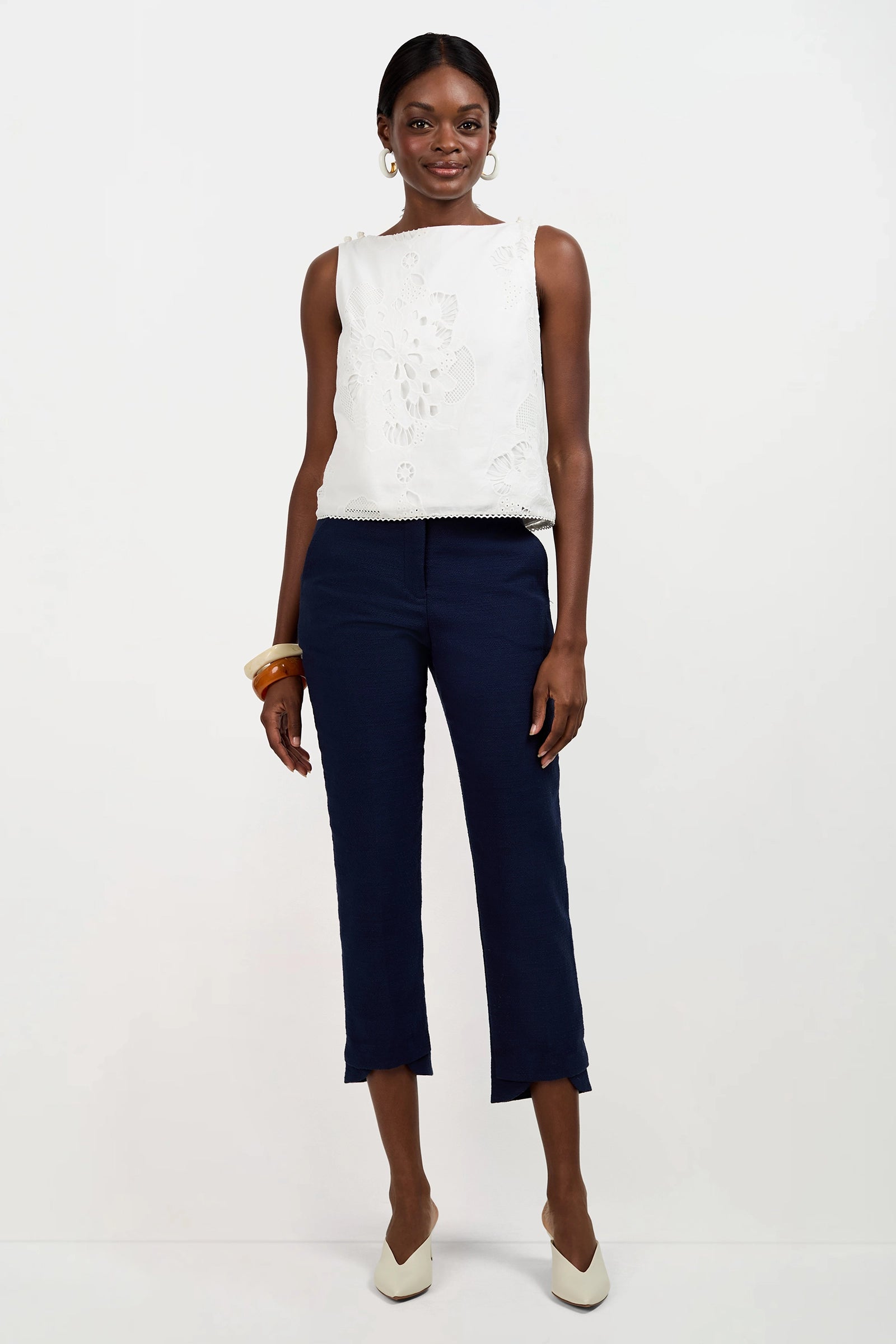 A woman stands against a plain white background wearing the sleeveless Alessandra Top with floral embroidery, paired with navy cropped pants, white heels, hoop earrings, and chunky bracelets. She smiles slightly with her hands at her sides.