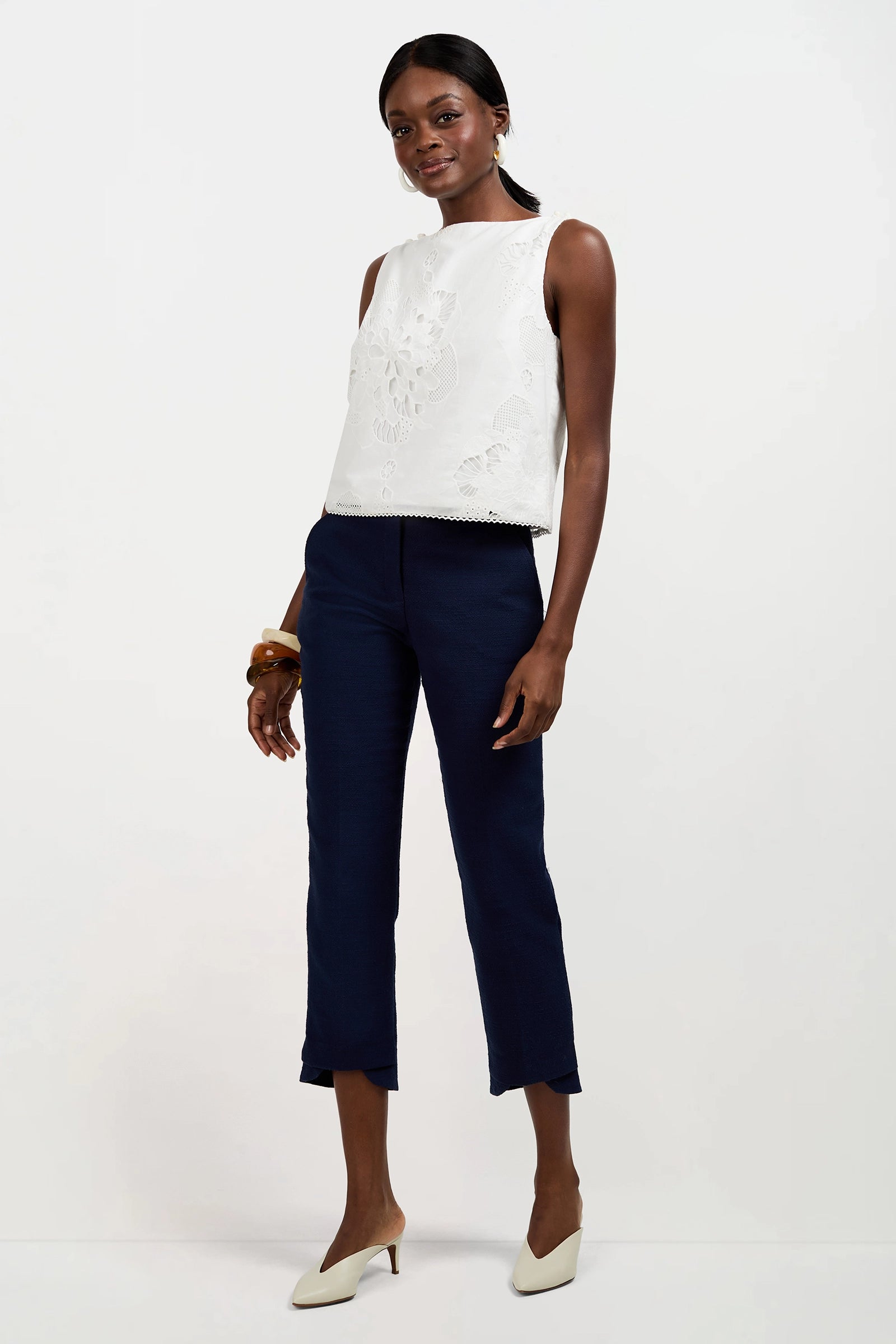 A woman poses against a white background wearing the sleeveless white embroidered Alessandra Top, navy cropped pants, beige heels, and a chunky brown bracelet. She stands with one leg bent and smiles gently.