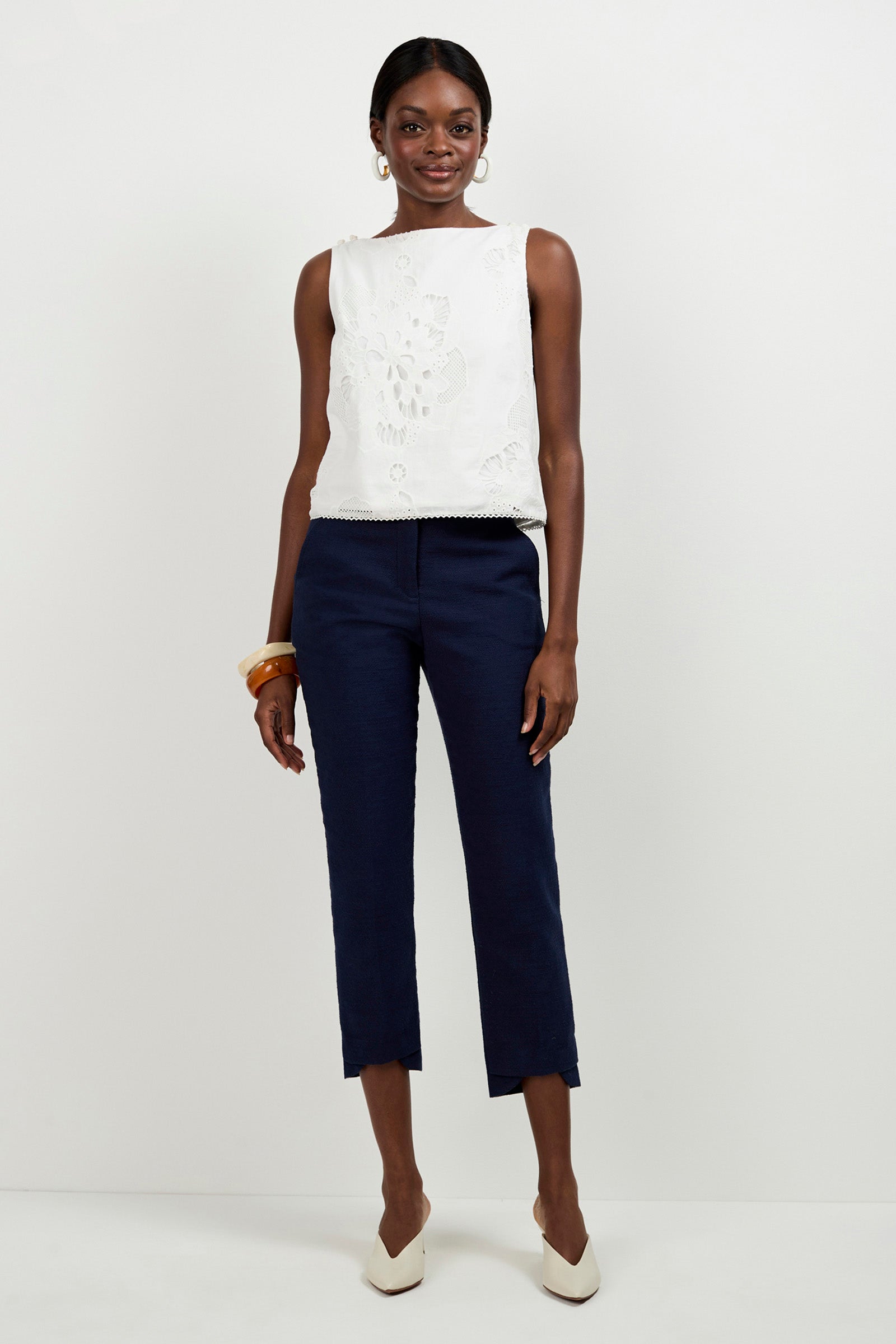 A woman stands against a plain white background, smiling as she wears the sleeveless white embroidered Alessandra Top, navy ankle-length pants, white pointed-toe heels, gold hoop earrings, and chunky bangles, her left hand relaxed by her side.