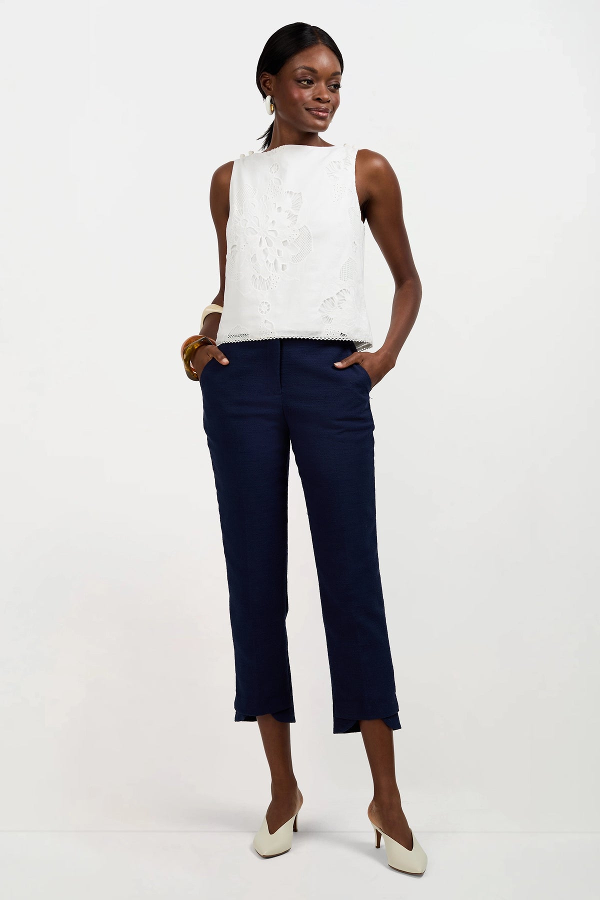A woman stands against a white background wearing the sleeveless Alessandra Top with floral cutouts, paired with navy ankle-length pants, white heels, and gold jewelry. She has her hands in her pockets and looks to the side.