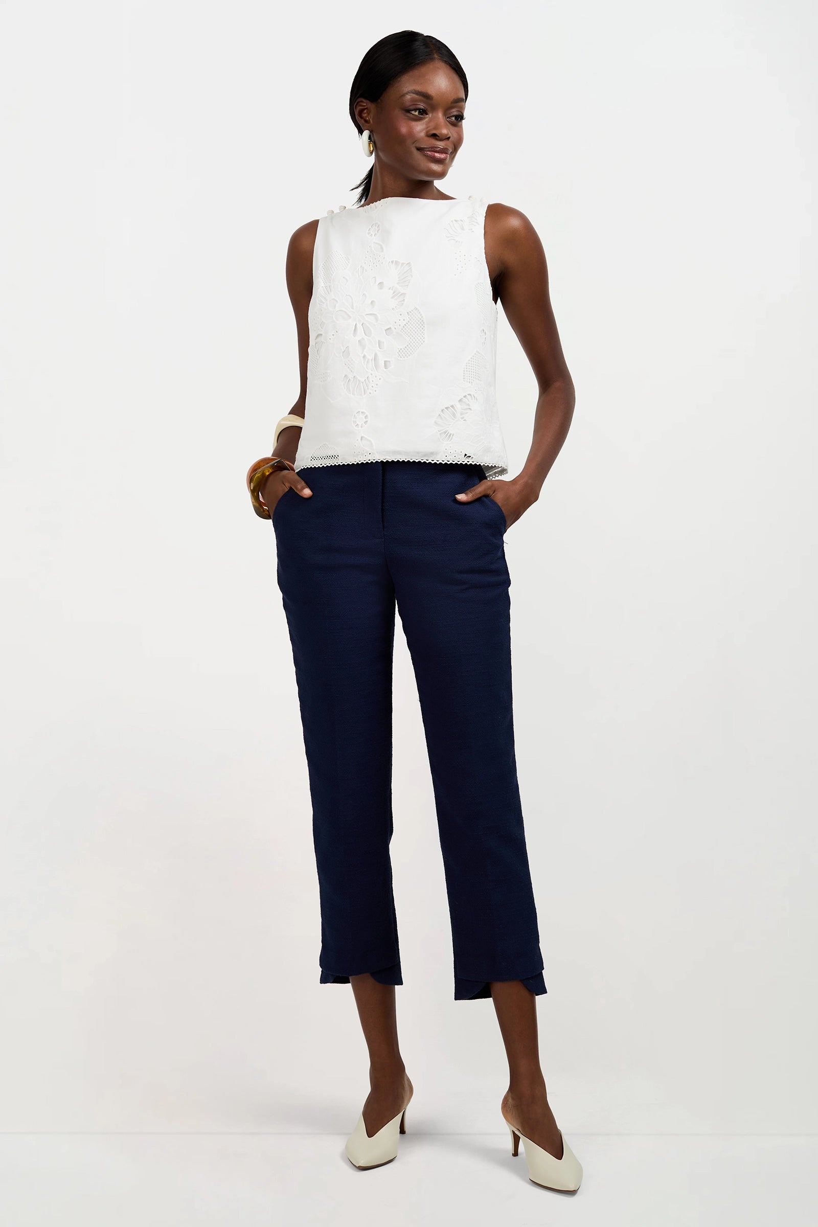 A woman stands against a white background wearing the sleeveless Alessandra Top with floral cutouts, paired with navy ankle-length pants, white heels, and gold jewelry. She has her hands in her pockets and looks to the side.