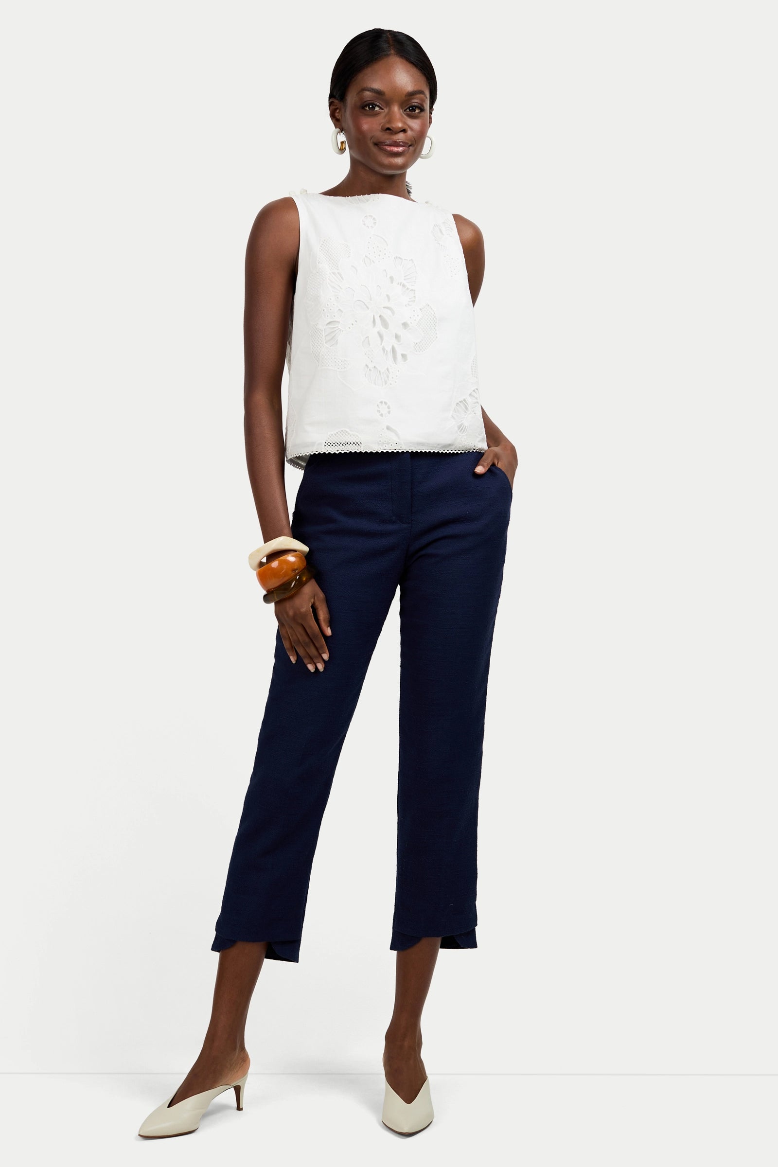 A woman stands confidently wearing the Alessandra Top, a sleeveless white top with floral detailing, paired with navy cropped pants, white heels, hoop earrings, and chunky bangles. Her hands are in her pockets as she smiles slightly.