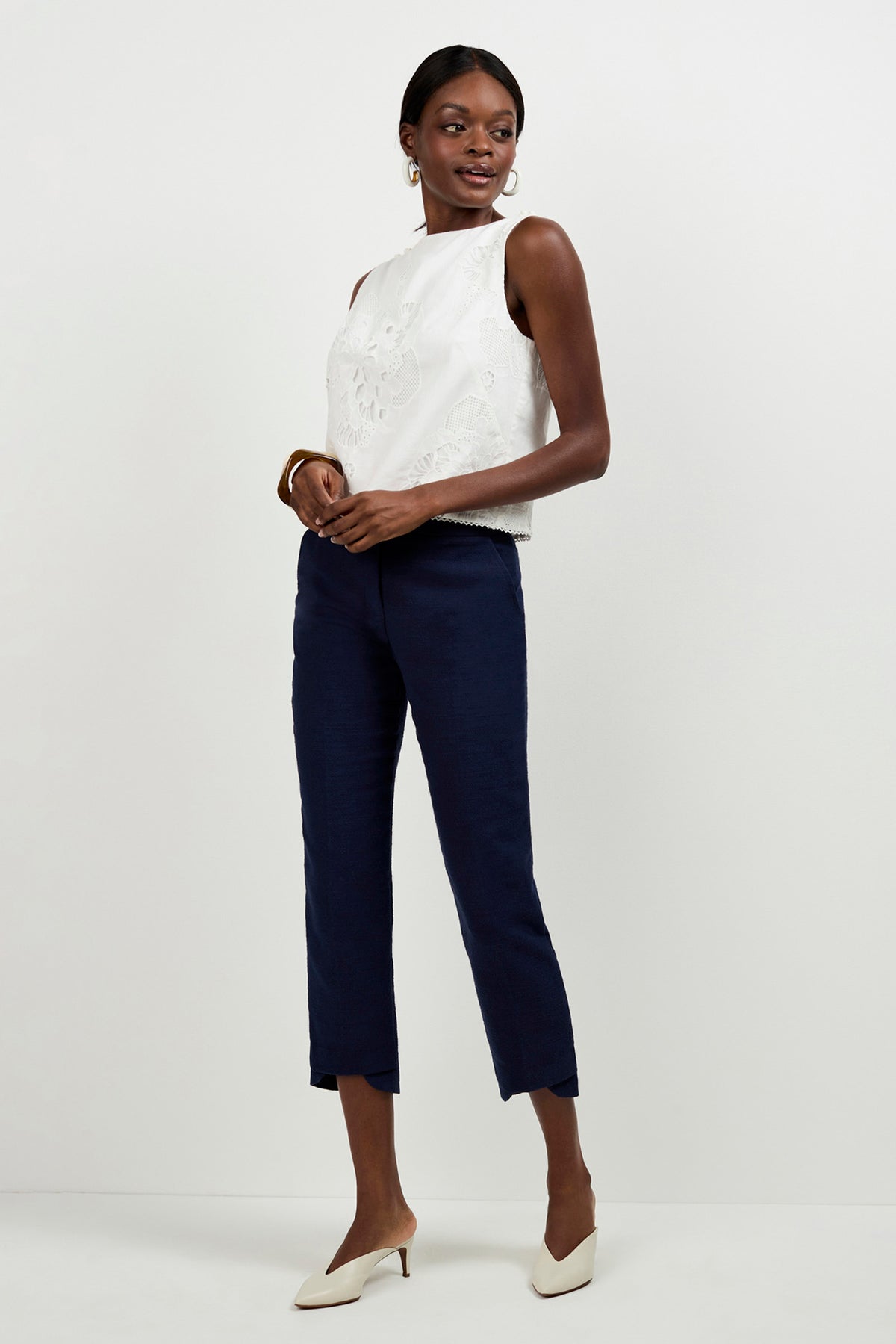 A woman smiles slightly and looks to the side against a white background, wearing Diffuse Pants in navy blue, a sleeveless white top with floral embroidery, white heeled shoes, hoop earrings, and a bracelet.