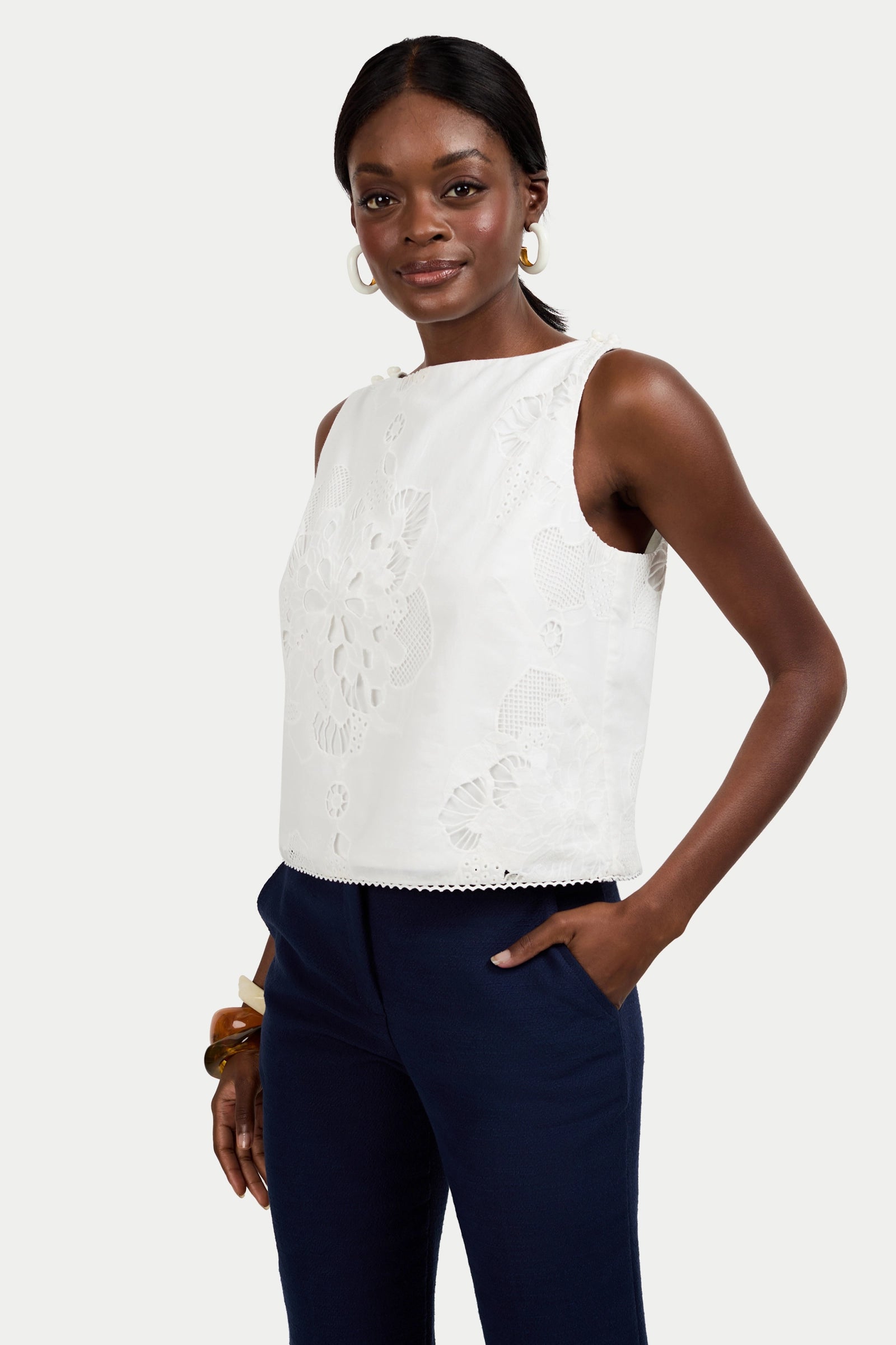A woman smiles softly against a plain backdrop, wearing the sleeveless white Alessandra Top with floral embroidery, navy pants, hoop earrings, and a bracelet. Her hair is pulled back and one hand rests in her pocket.
